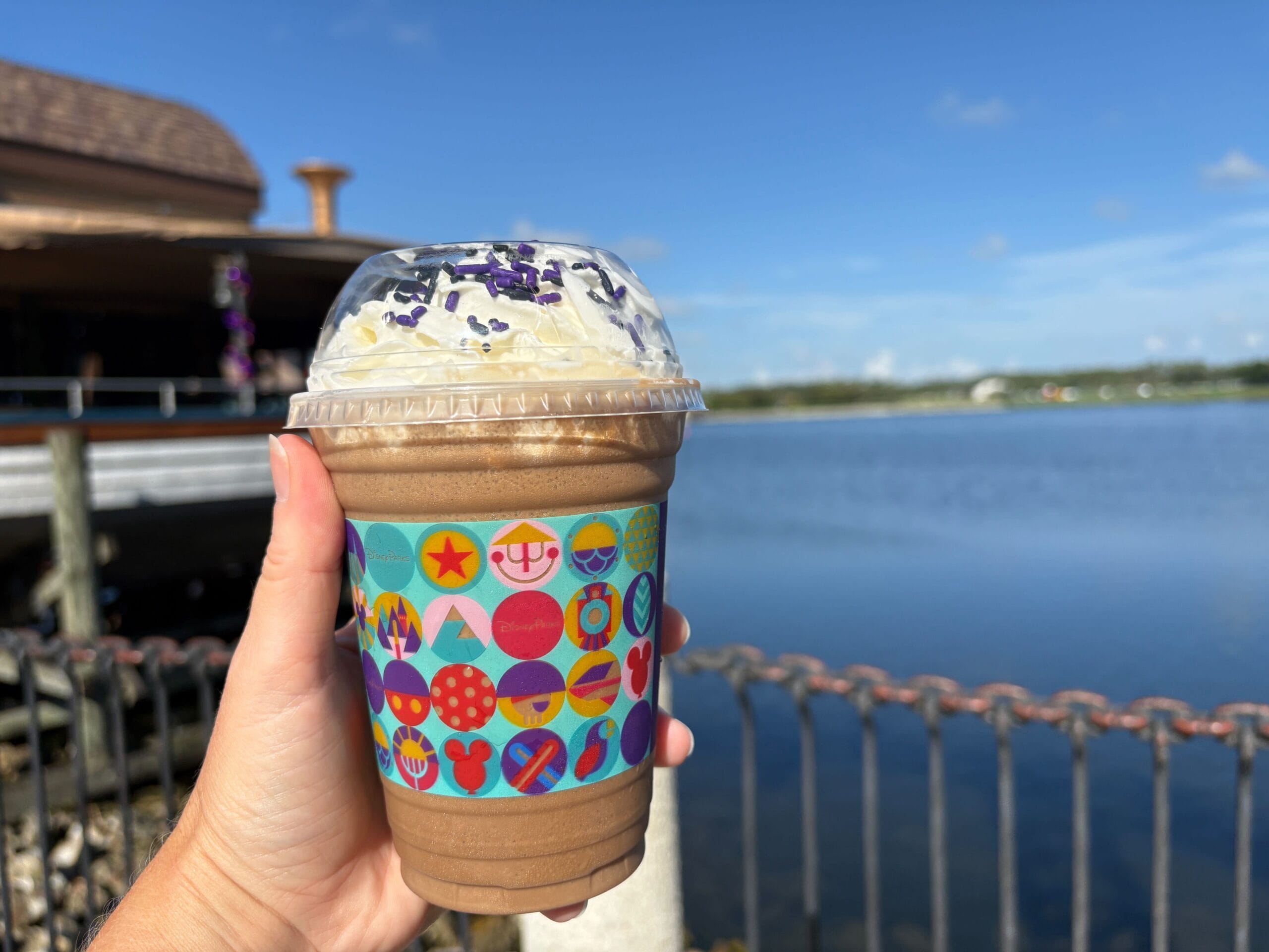 A hand holds a chocolate milkshake with whipped cream by a lake at Disney Springs on a sunny day.