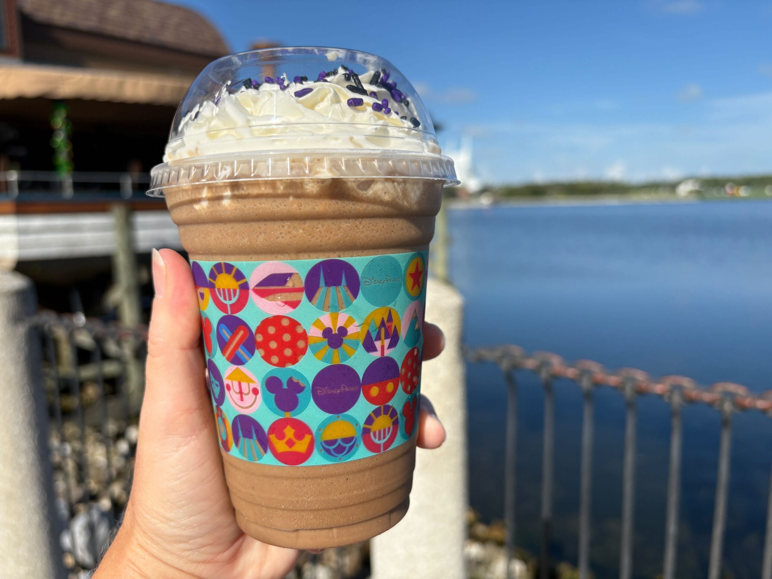 A hand holds a chocolate milkshake with whipped cream by a lake in Disney Springs, in a colorful Disney-themed cup.