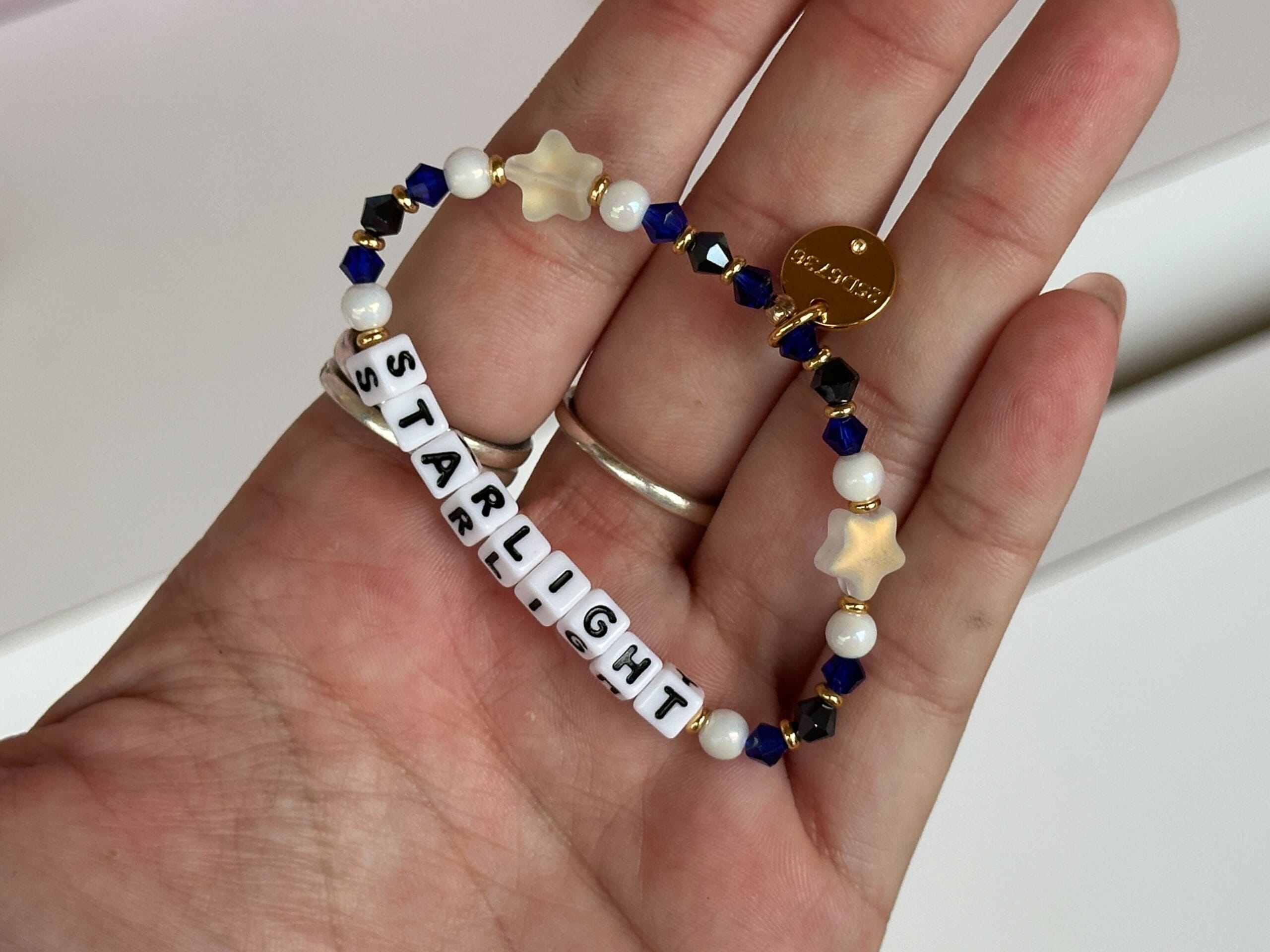 A hand holds a Starlight bracelet from Little Words Project with blue, white, and gold beads spelling "STARLIGHT.