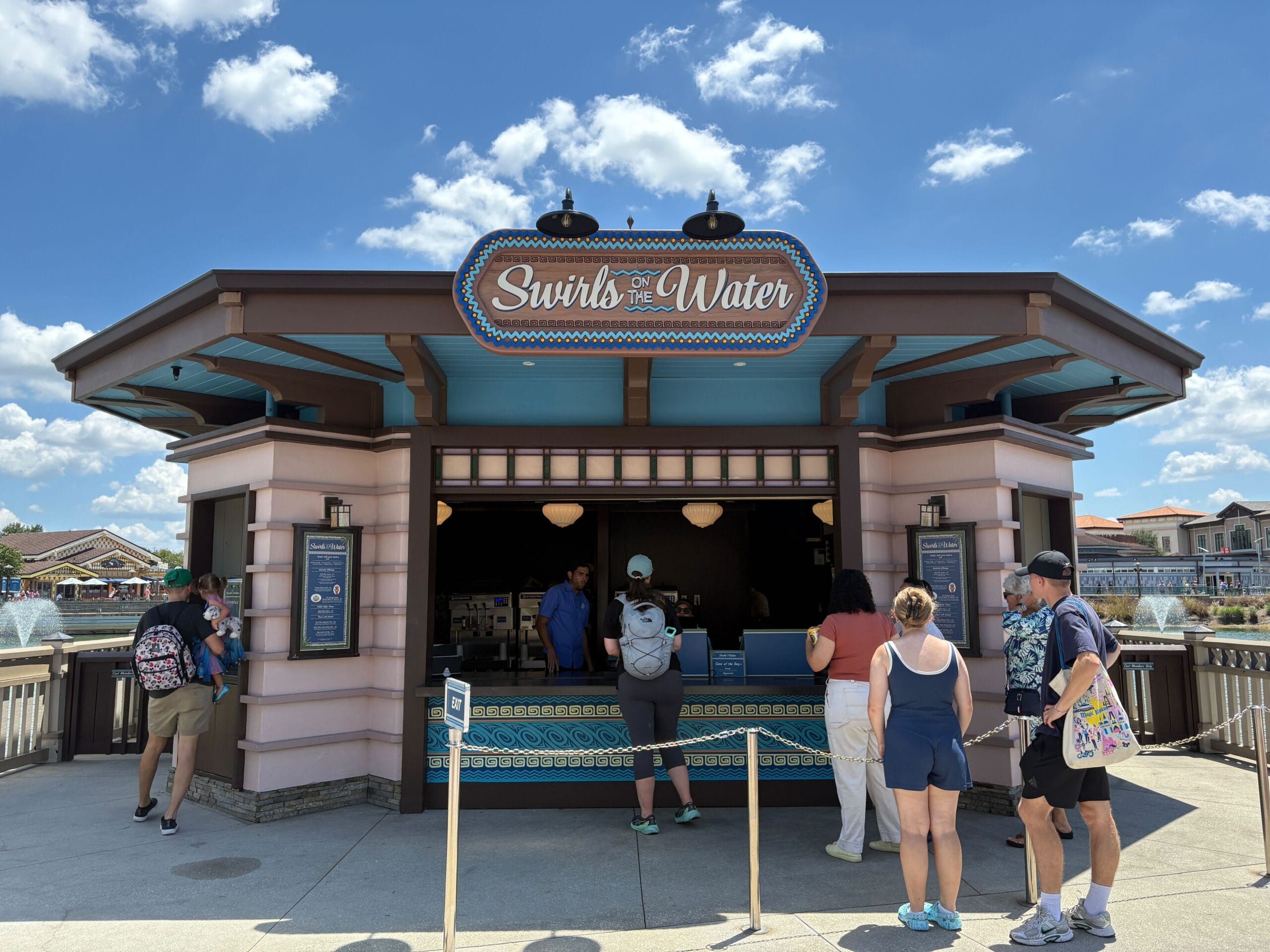 People line up at Disney Springs' Swirls on the Water kiosk for Halloween food on a sunny day with blue skies.