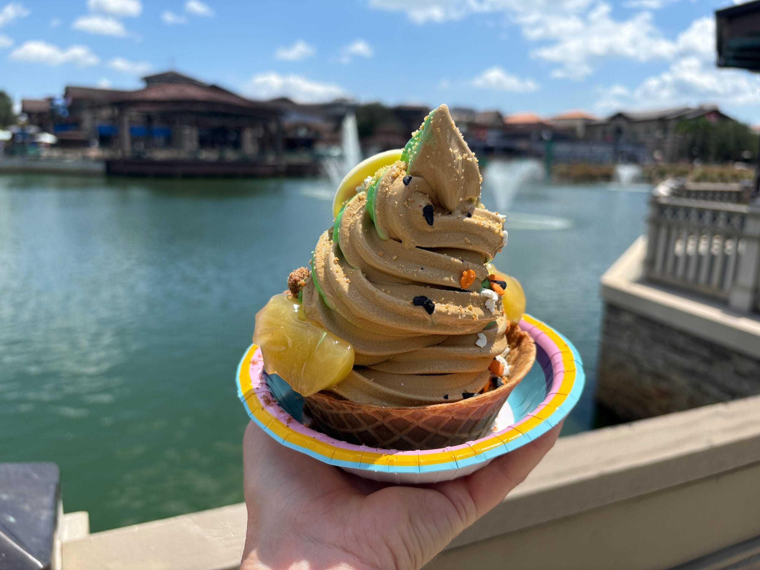 A hand holds Halloween Food soft serve ice cream in a waffle bowl with toppings, by Disney Springs on a sunny day.