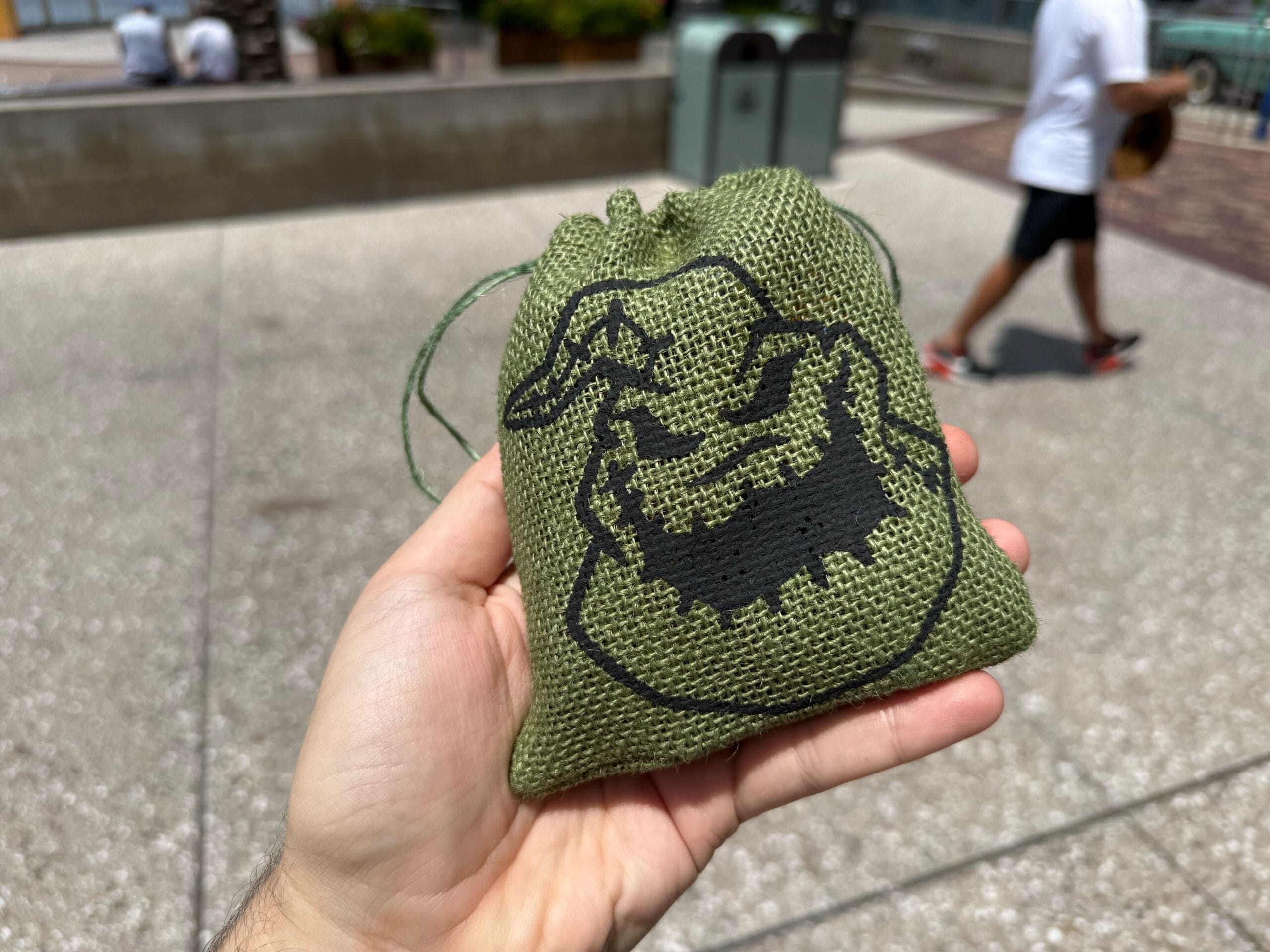 A hand holds a green drawstring bag with a black cartoon face, perfect for Halloween Food at Disney Springs.