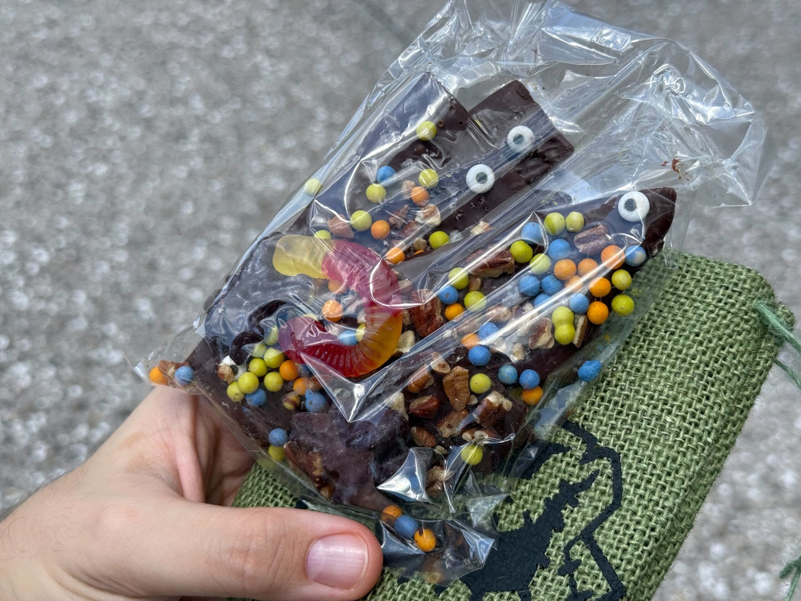 A hand holds a bag of Halloween food chocolate bark topped with candy eyes, gummy worms, and candy sprinkles.