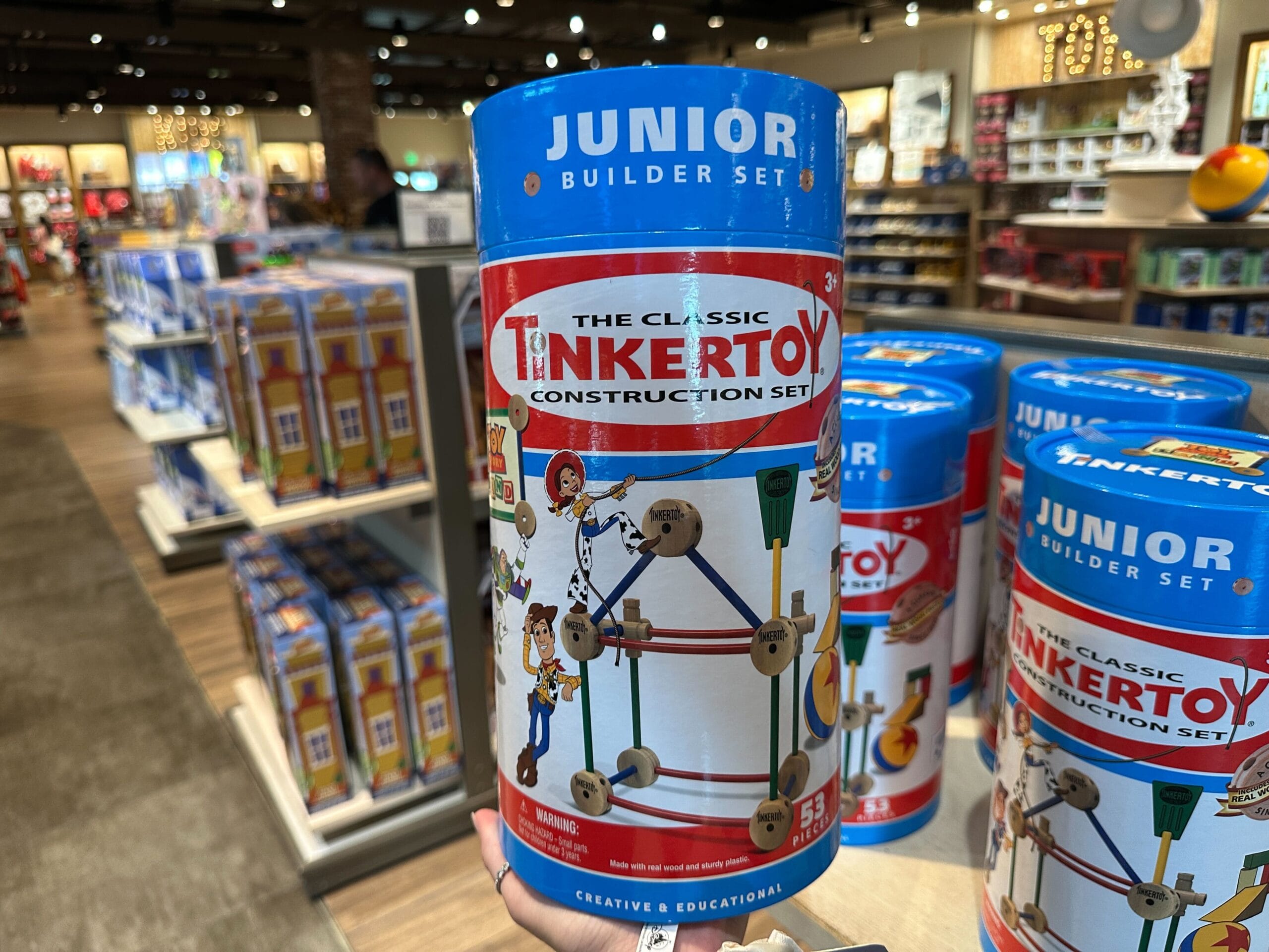 A hand holds a Tinker Toy Construction Set canister in a toy store filled with colorful displays.