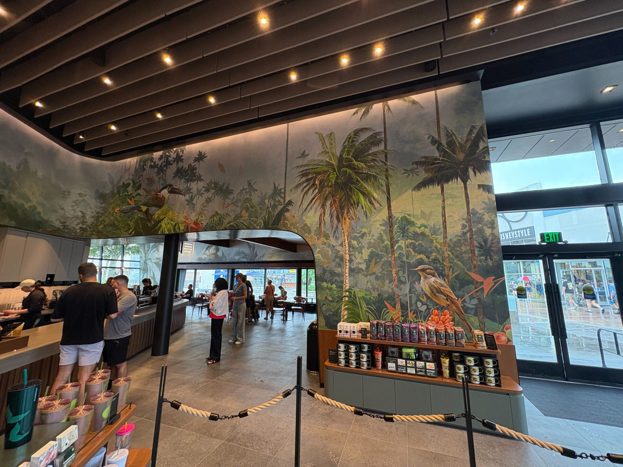 Coffee shop interior at Disney Springs with tropical mural, people ordering, and Starbucks merchandise on display.