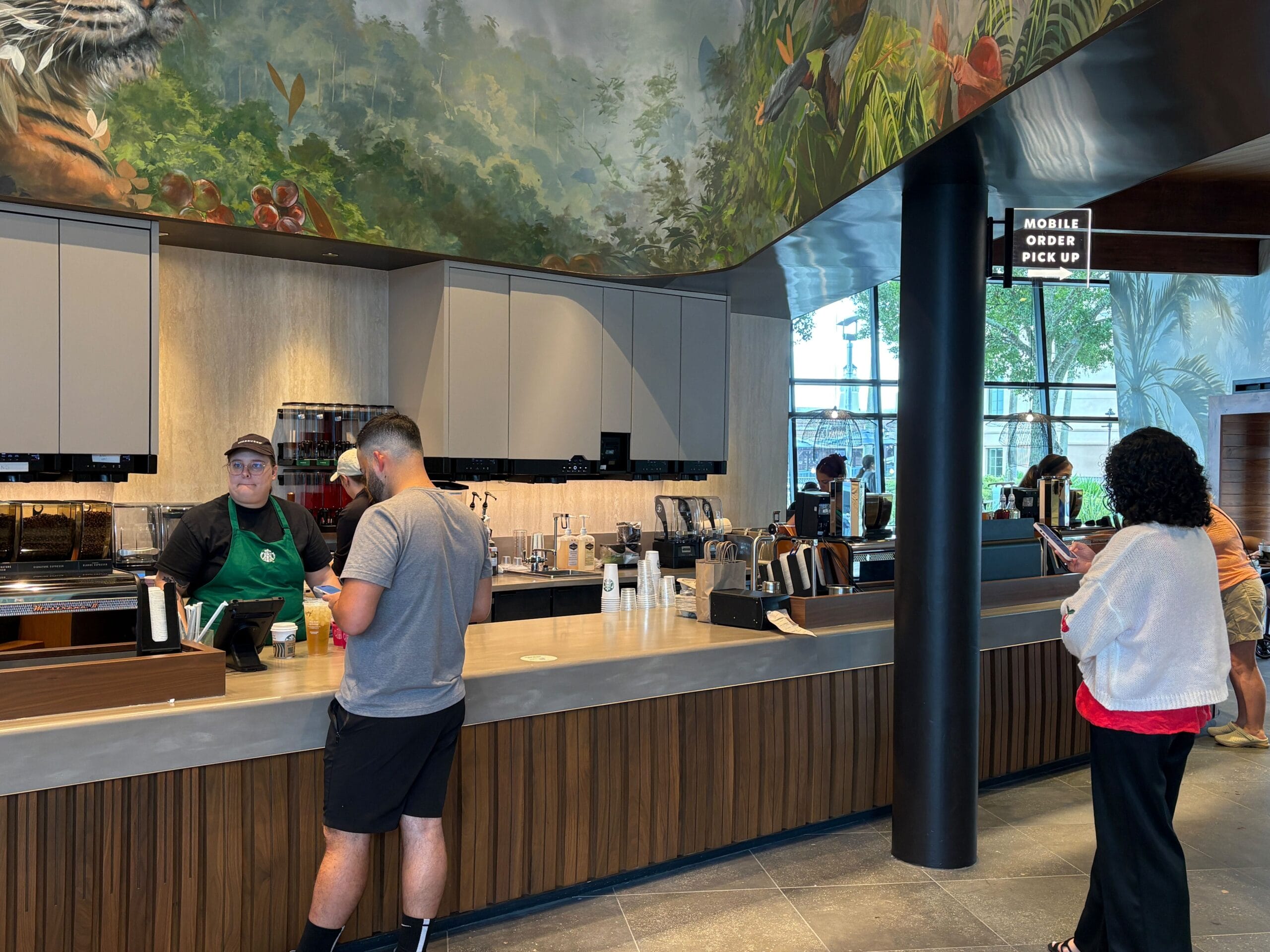 People wait at a counter in a modern Starbucks as it reopens at Disney Springs, with a jungle-themed mural and pick-up sign.