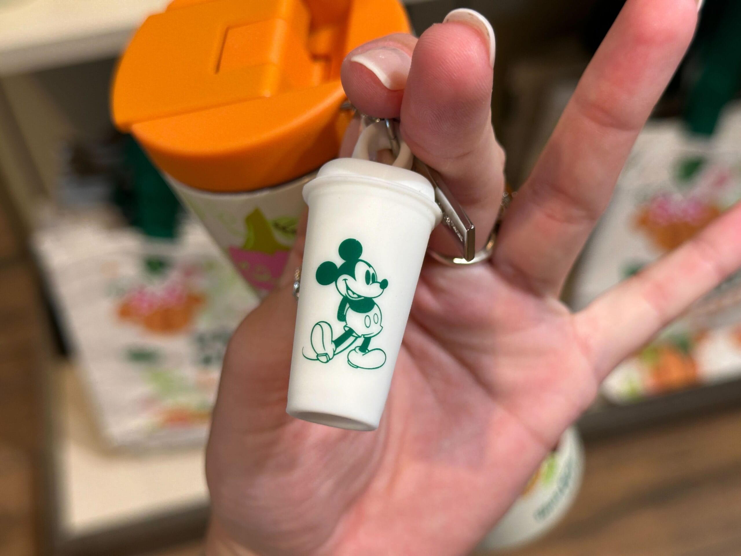 A hand holds a small white keychain cup with a green Mickey Mouse graphic, perfect for Disney Parks and Starbucks Pumpkin Spice fans.