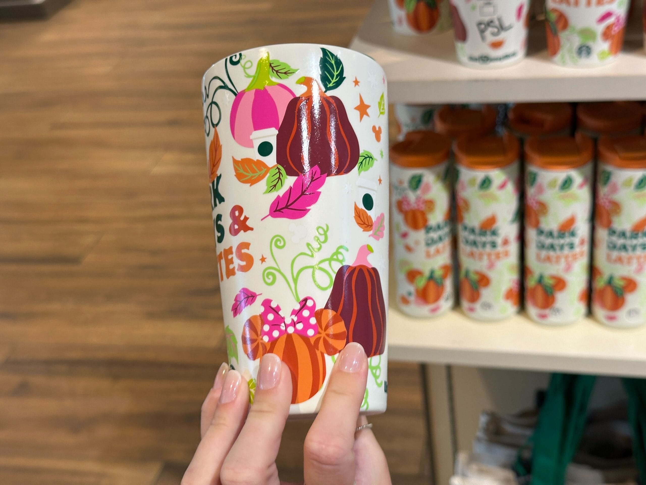 A hand holds a fall-themed Starbucks Pumpkin Spice cup with colorful pumpkins in front of similar cups on a shelf part of Park Days and Lattes collection