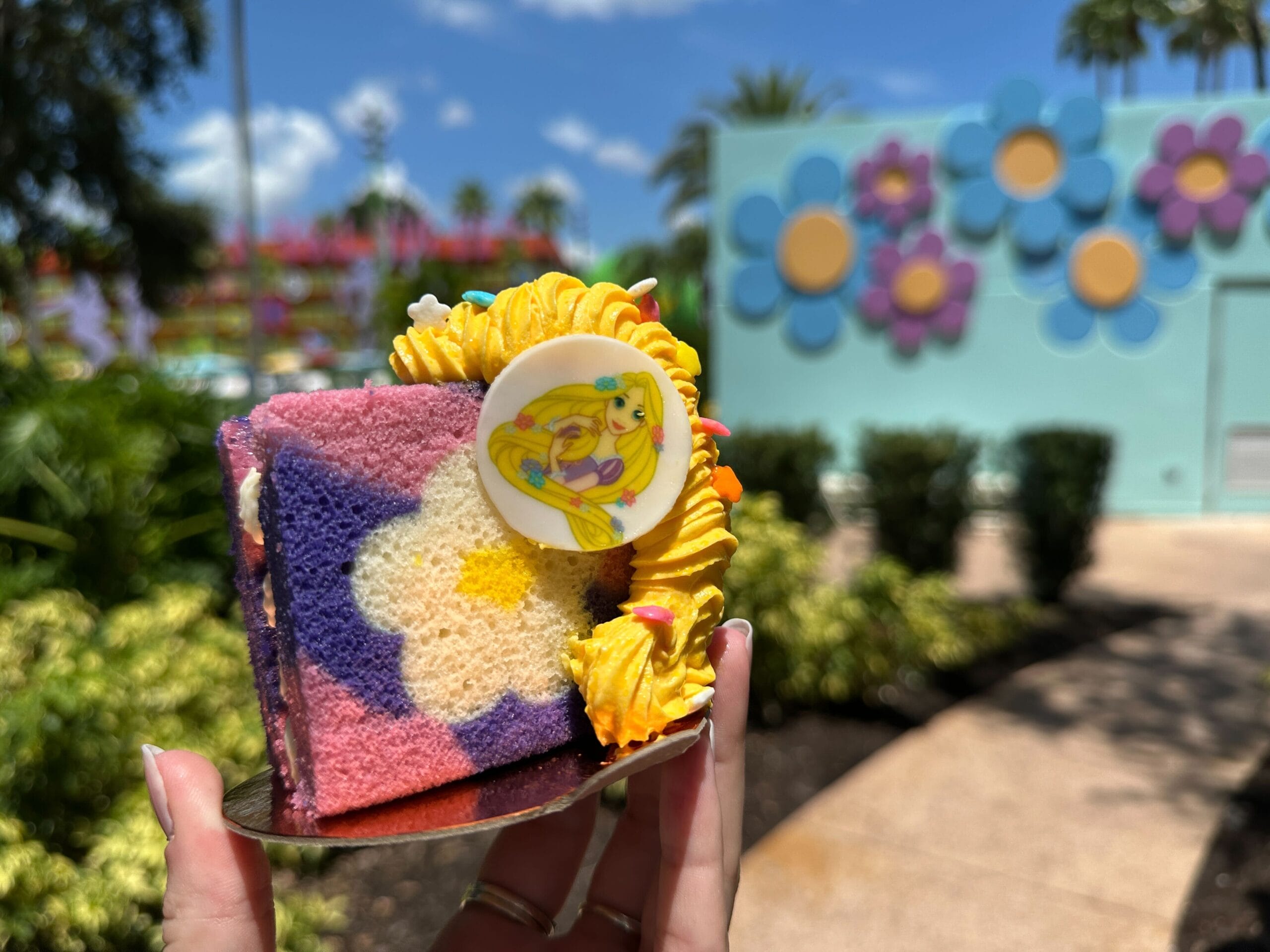 A hand holds the Rapunzel Berries and Cream Cake with a cartoon topper at Everything POP Dining, flower mural behind.