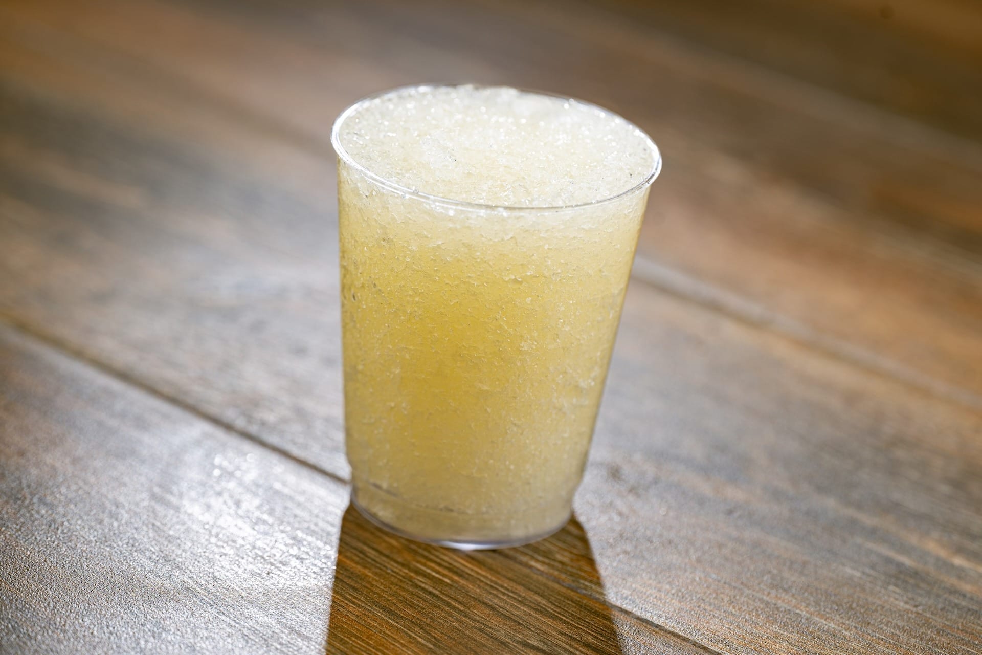 A clear plastic cup filled with a light yellow slushy drink at the EPCOT Food & Wine Festival on a wooden surface.