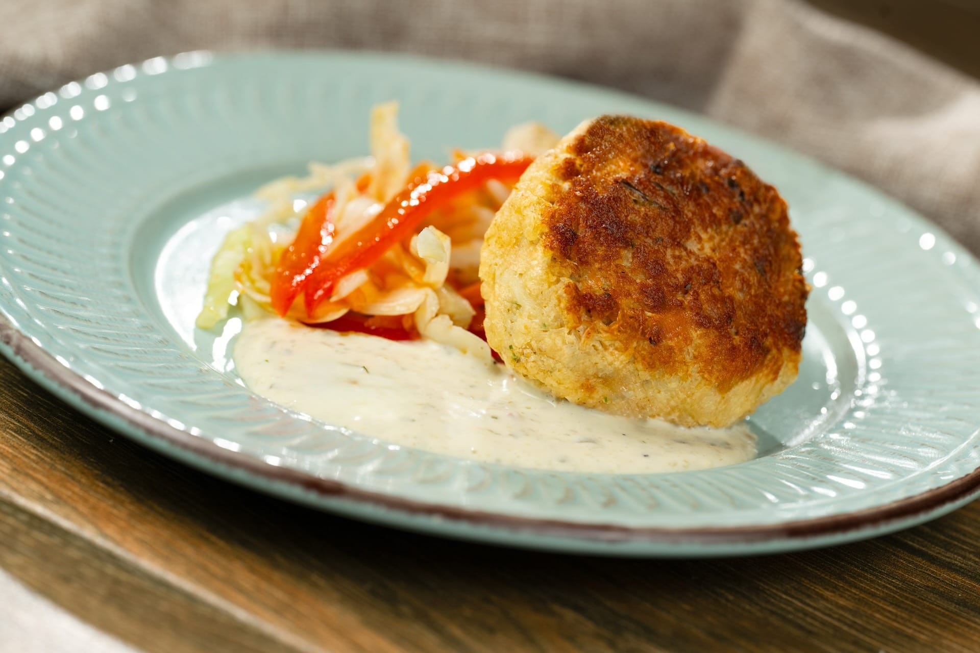 A golden-brown fish cake on creamy sauce, featured in EPCOT Food & Wine Festival 2025 menus with sautéed cabbage and peppers.