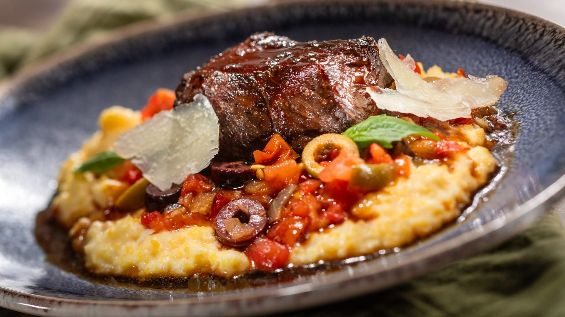 A braised beef steak on creamy polenta, featured on the 2025 menus at EPCOT Food & Wine Festival, garnished with basil.