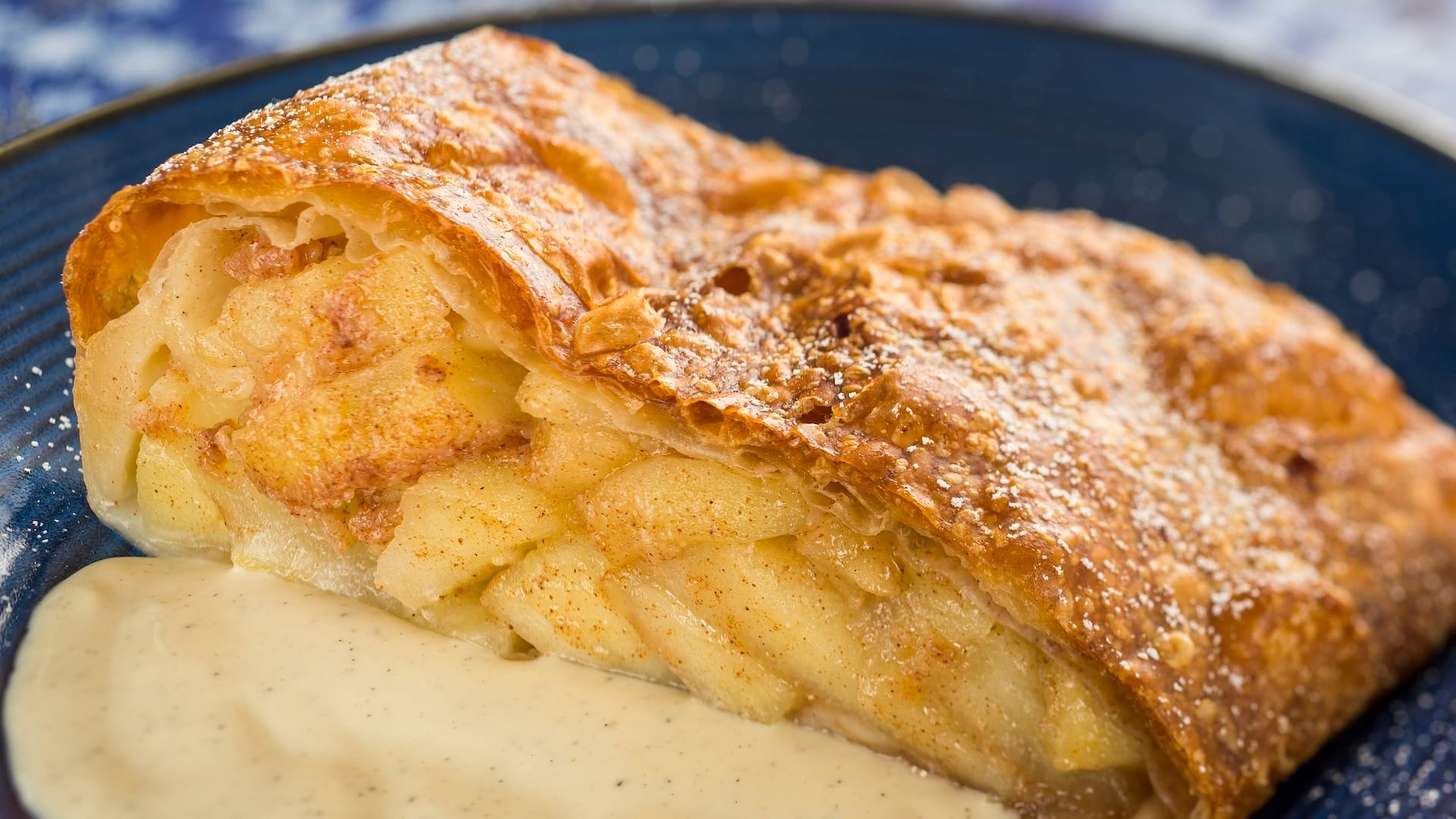 A slice of apple strudel with golden crust, from the EPCOT Food & Wine Festival 2025 menus, served with vanilla sauce.