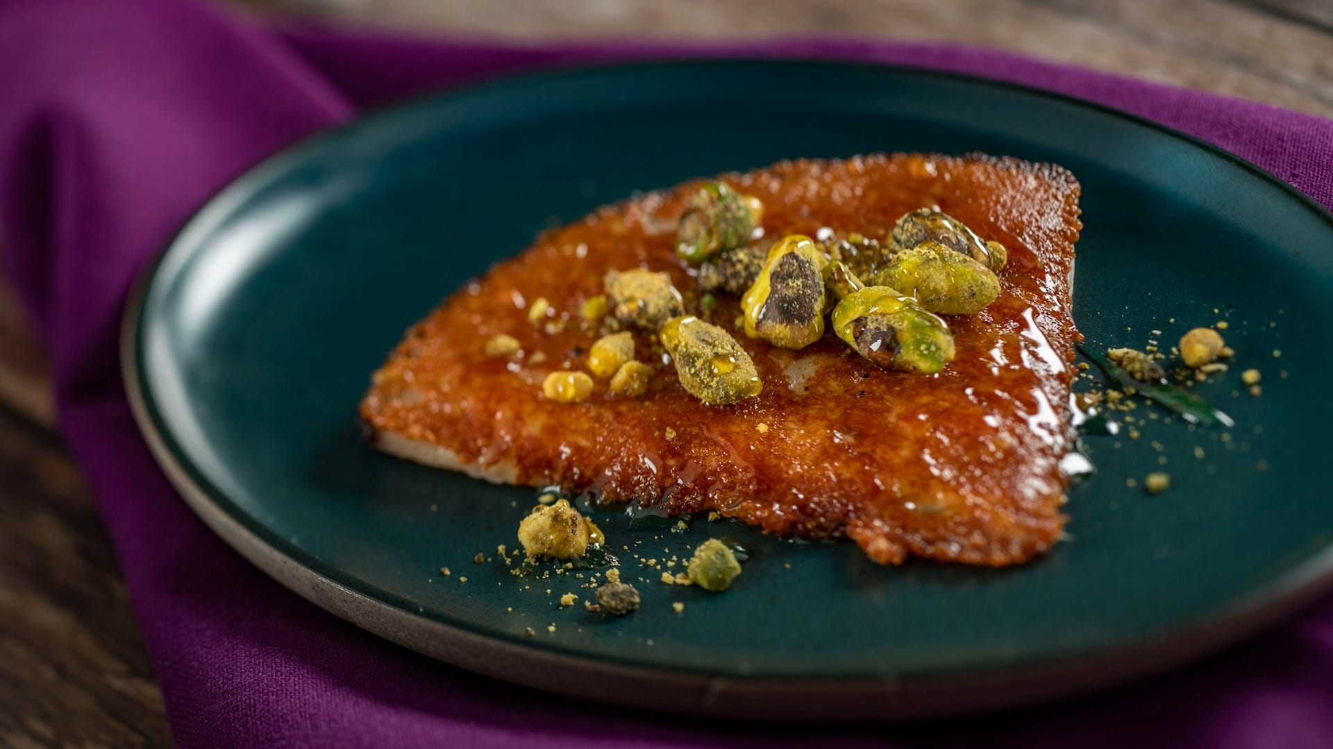 A triangular piece of brown dessert topped with pistachios at the EPCOT Food & Wine Festival on a dark plate.