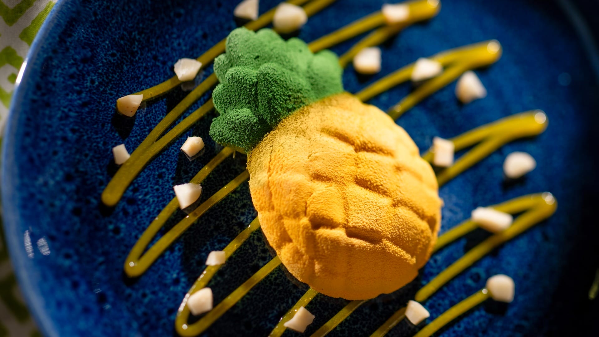 A pineapple-shaped dessert from EPCOT Food & Wine Festival 2025 on a blue plate with yellow sauce and white cubes.