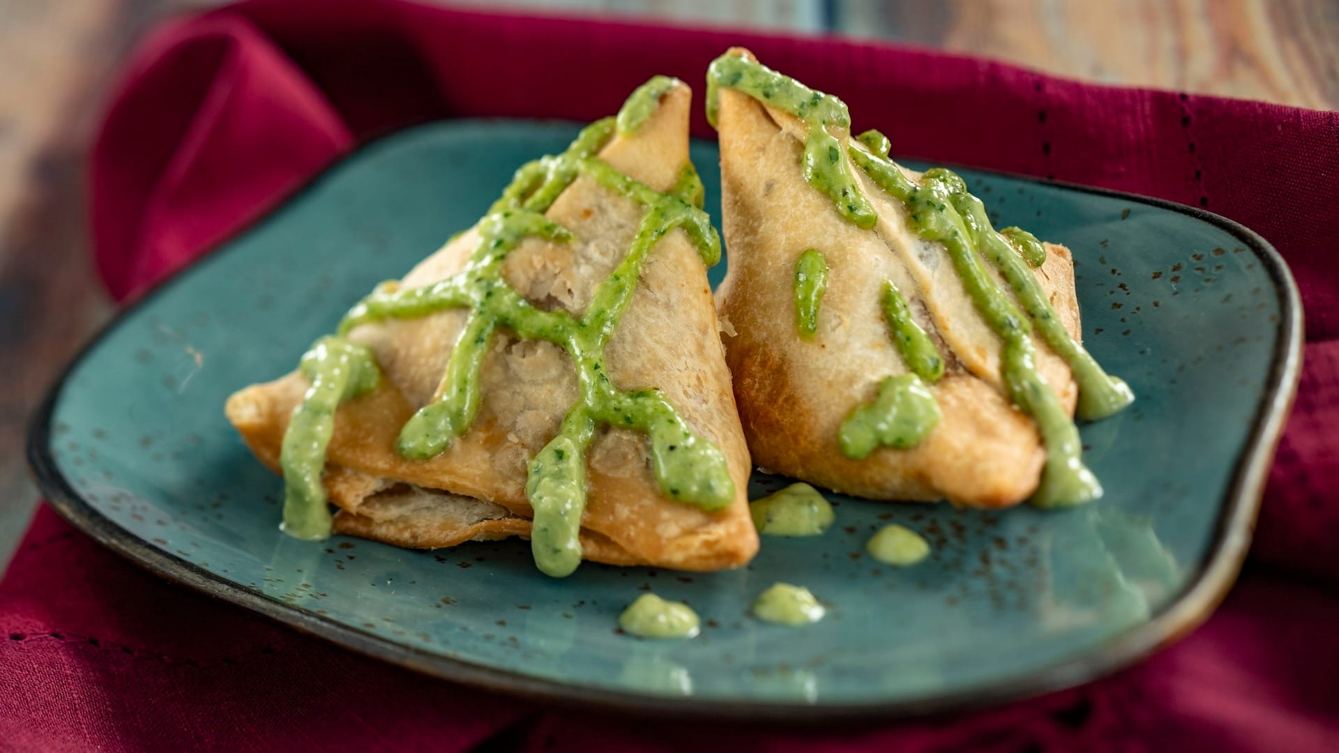 Two golden-brown samosas with green sauce, inspired by EPCOT Food & Wine Festival, served on a blue plate.