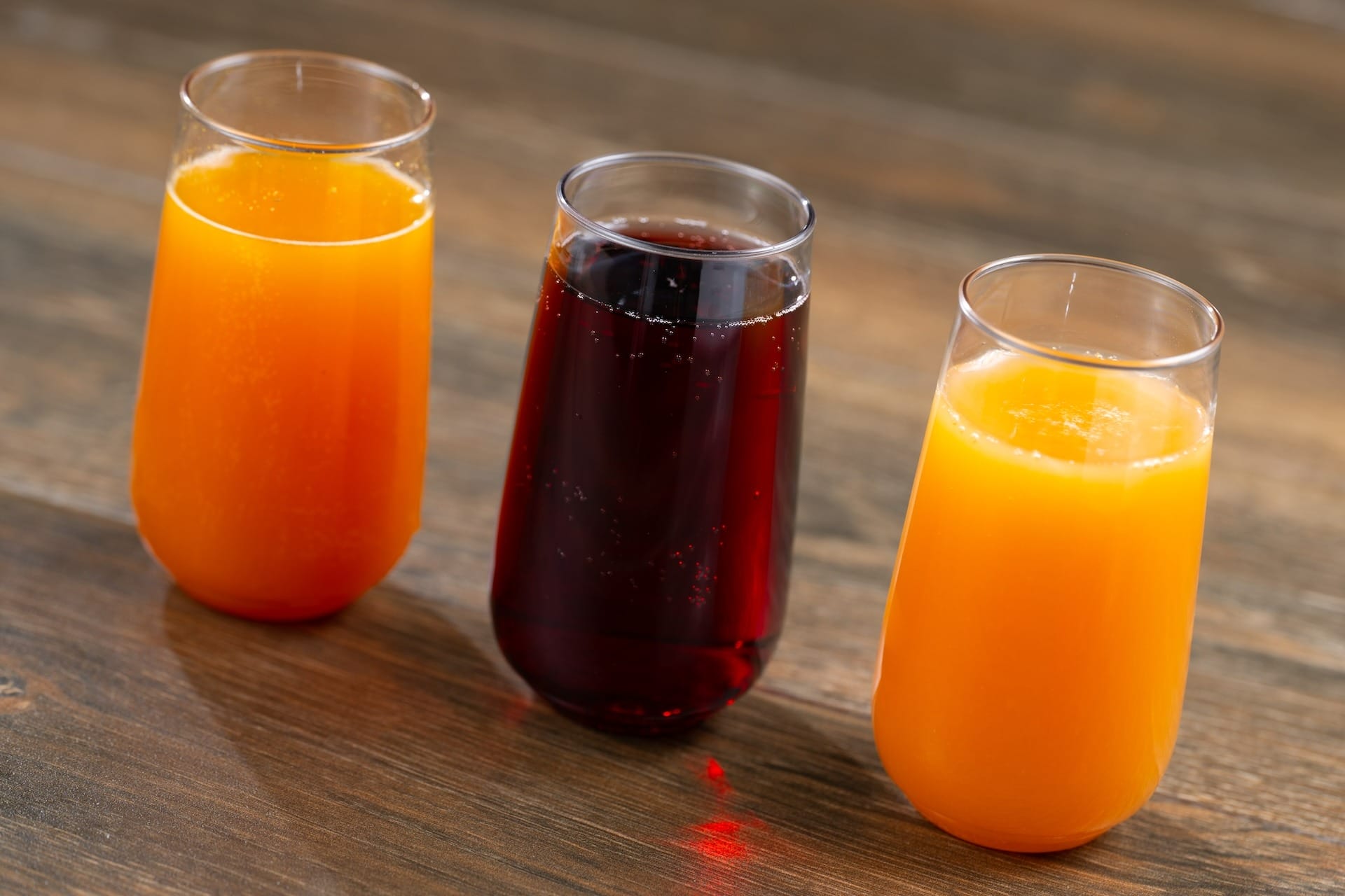 Three glasses—two with orange juice, one dark red—from the EPCOT Food & Wine Festival sit on a wooden surface.
