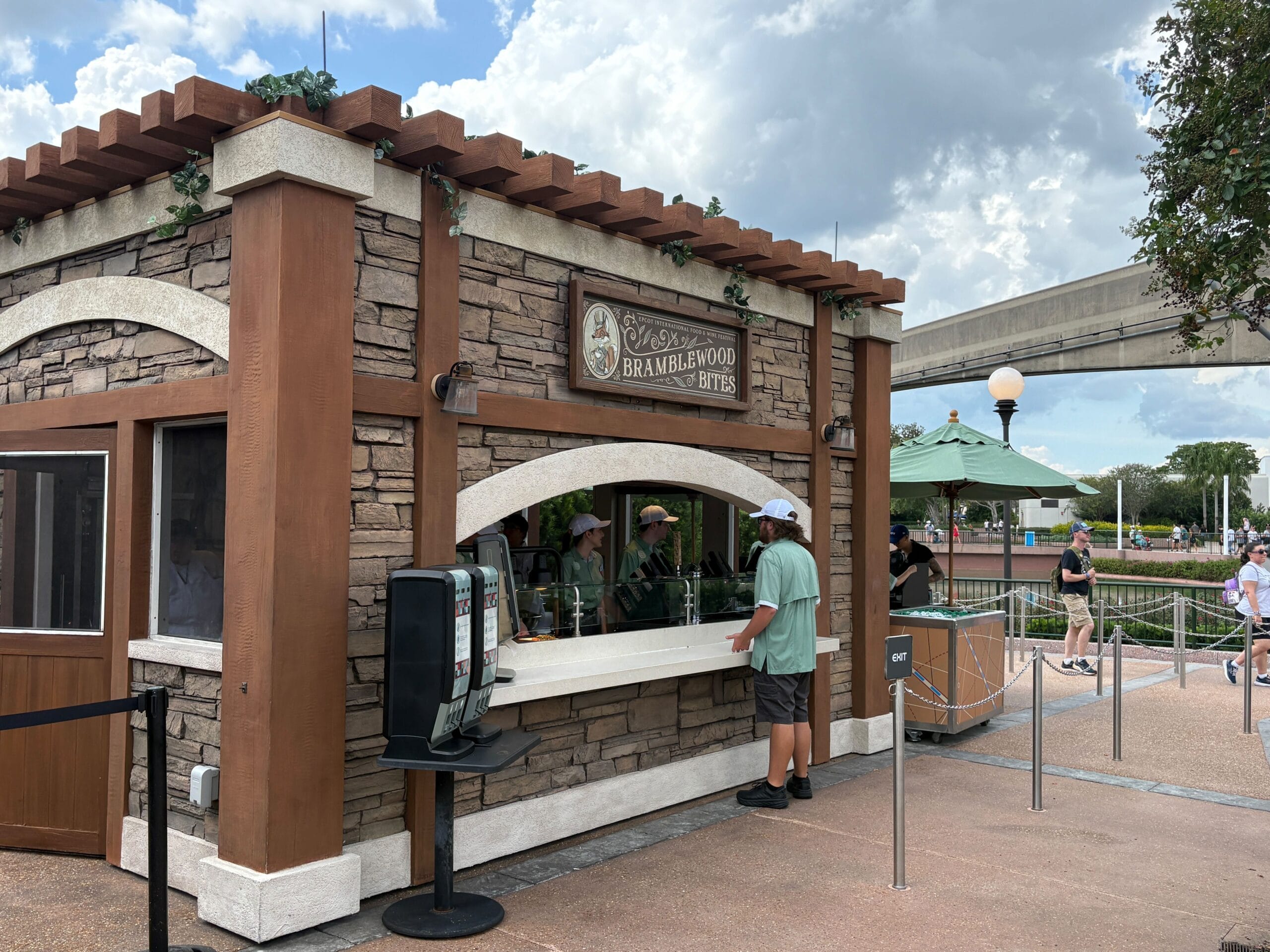 A man orders New Food & Wine at a rustic outdoor booth labeled "Bramblewood Bites" in a park setting.