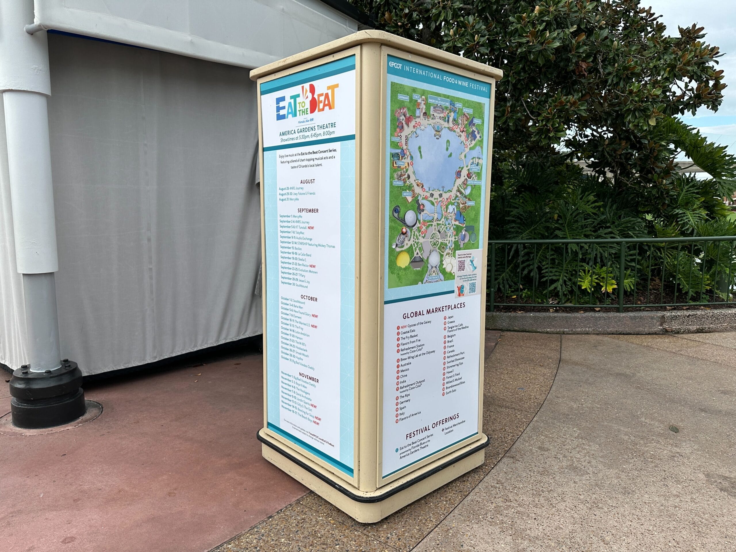 Large festival guide with a map and event details stands on a sidewalk near greenery and a white structure.