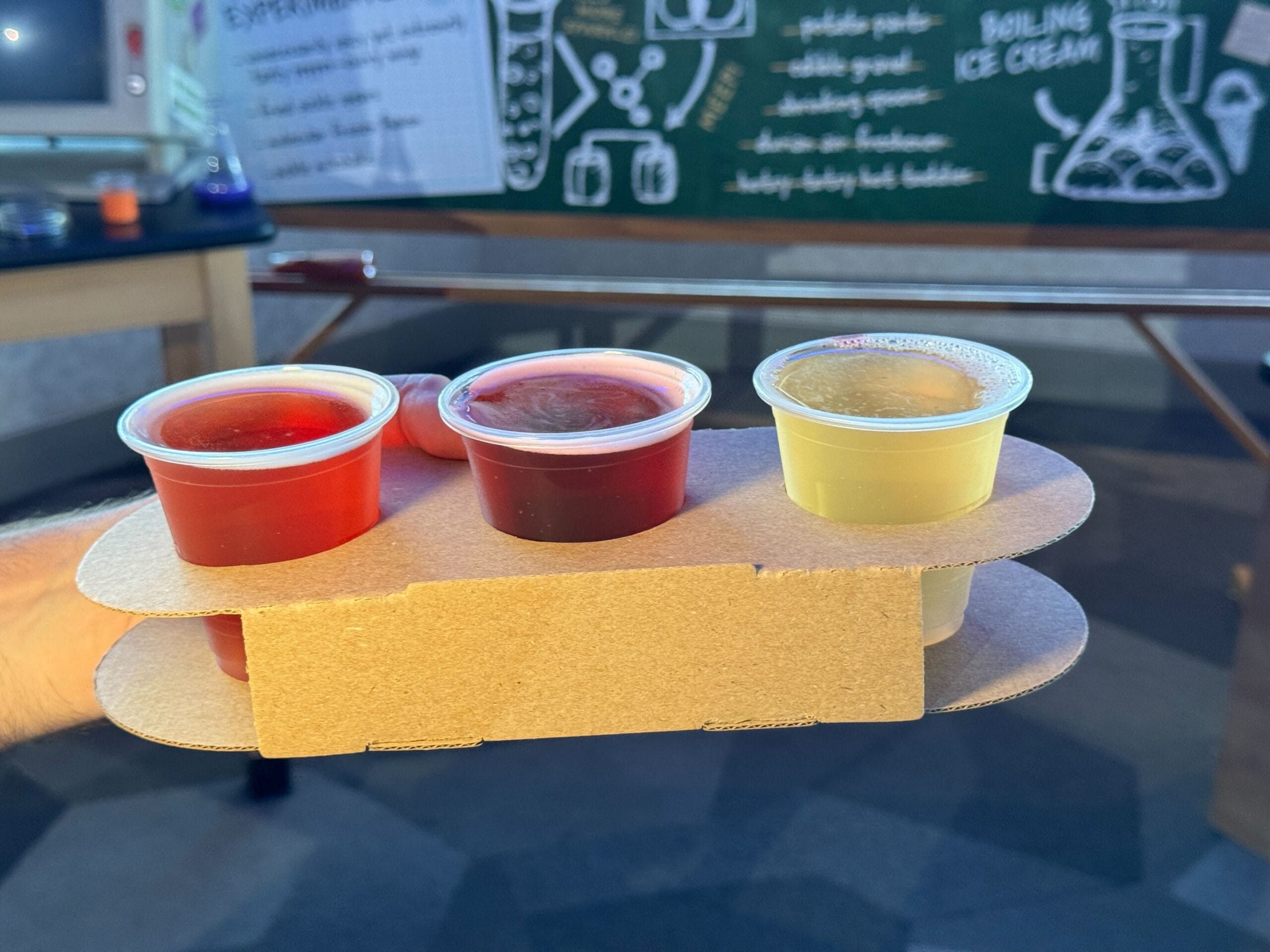 A hand holds a cardboard tray with three small cups of New Food & Wine in front of a chalkboard with science drawings.