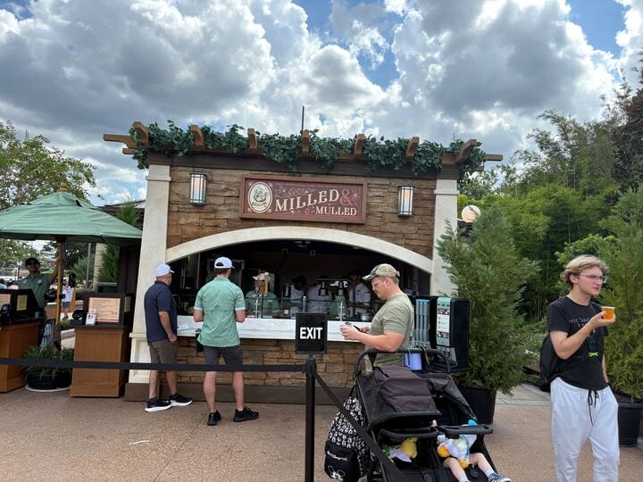 People gather at the "Milled & Mulled" New Food & Wine booth outdoors, with greenery and a stroller nearby.