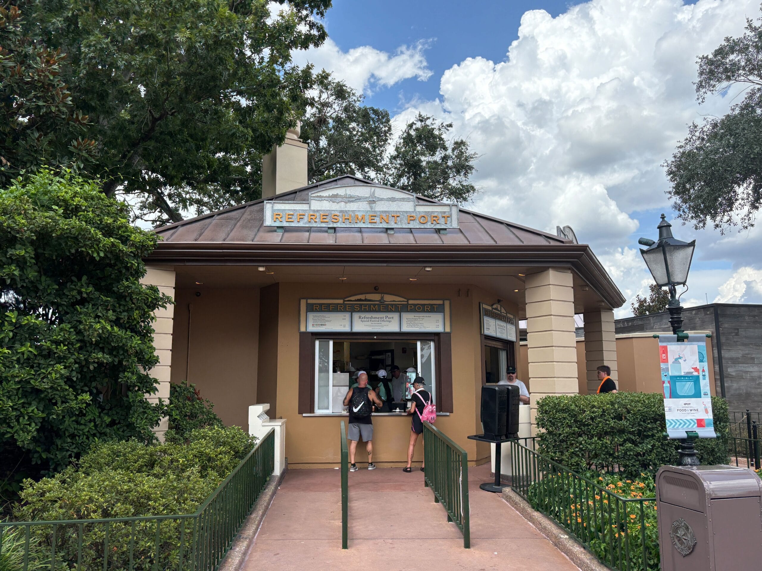 People standing at the Refreshment Port counter, sampling New Food & Wine, with trees and a cloudy sky in the background.