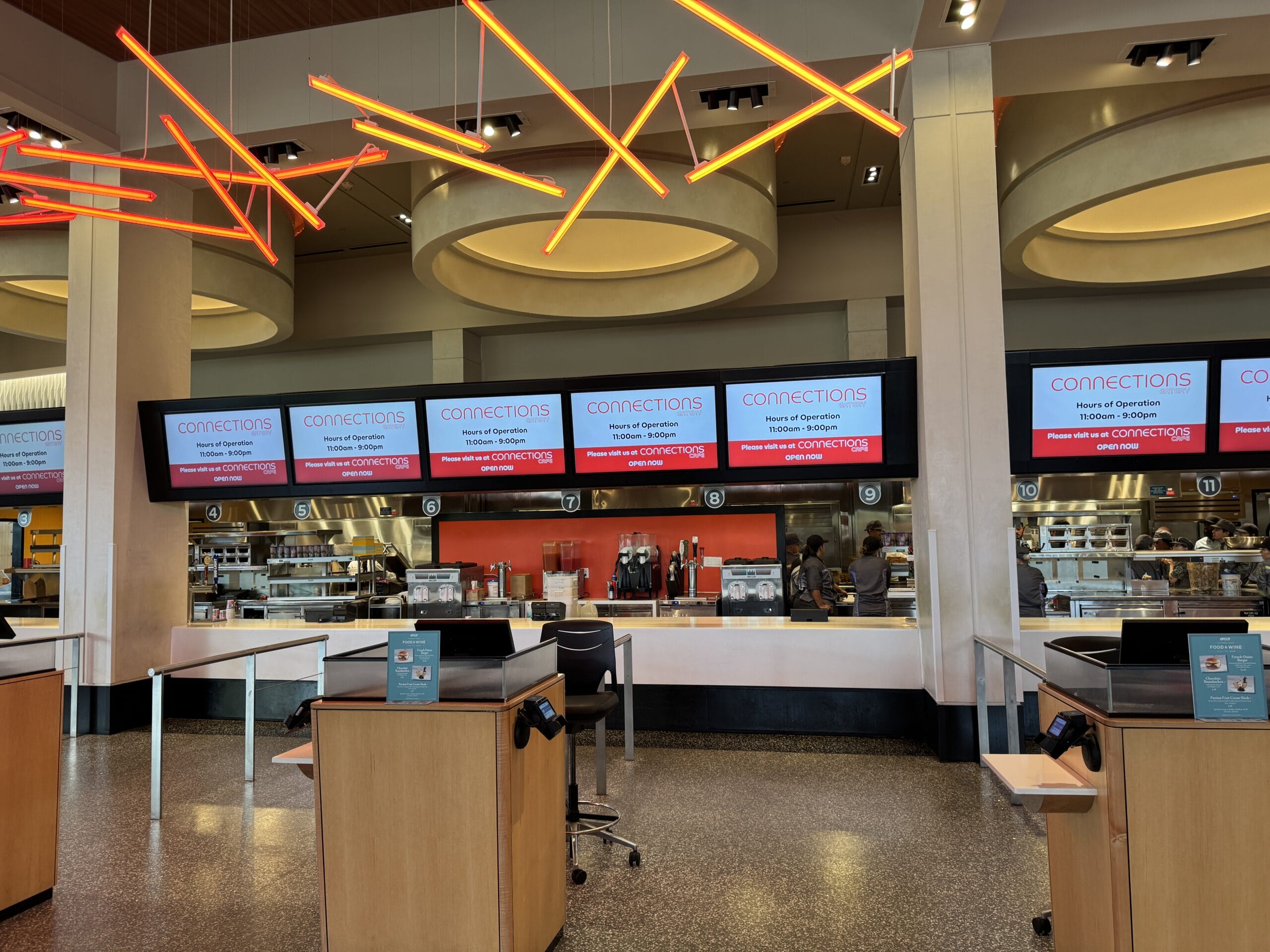 Modern cafeteria with New Food & Wine vibes—bright orange lights, digital menus, and a kitchen view in the background.