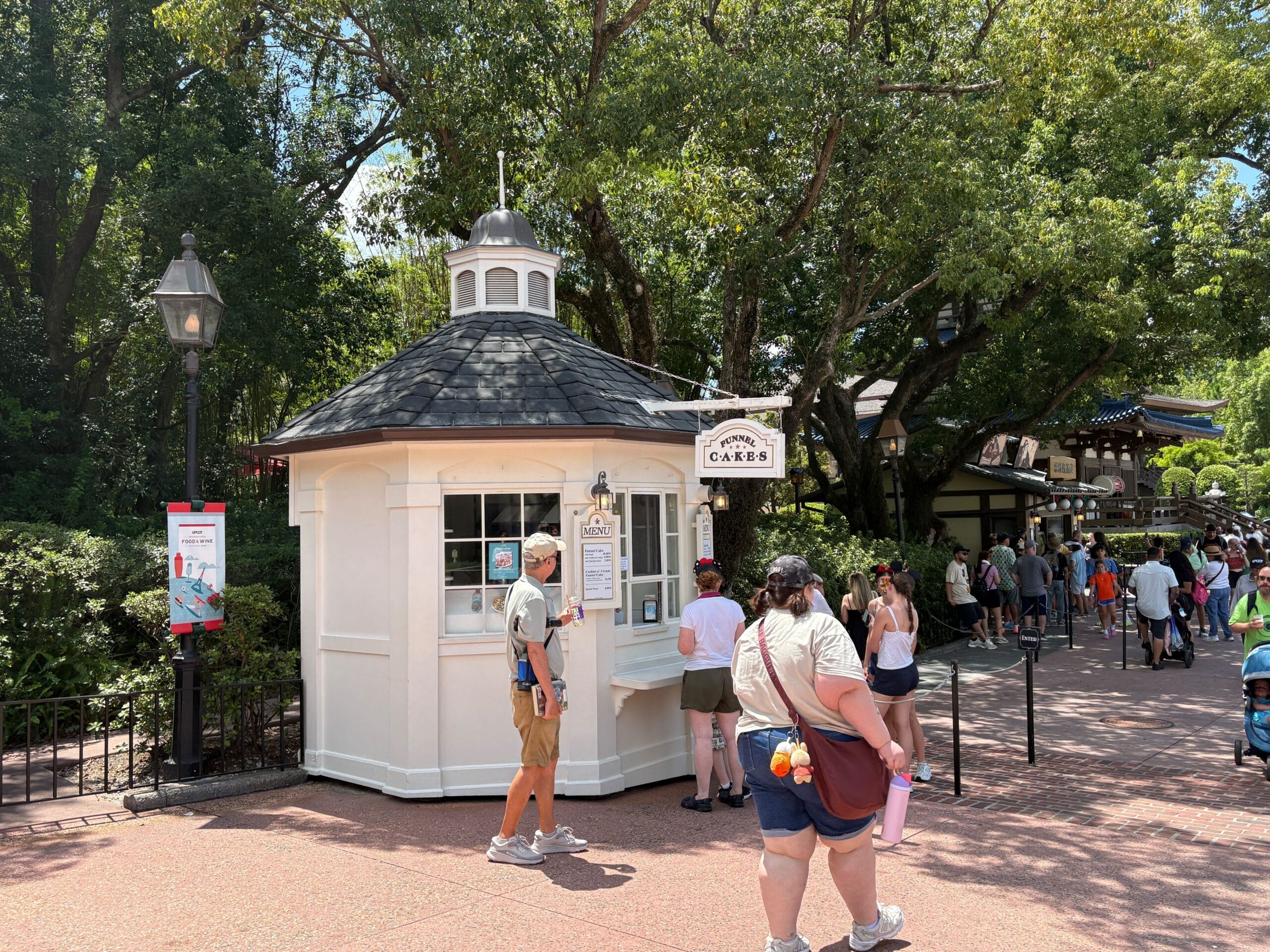 People stand near a small white outdoor kiosk serving New Food & Wine, surrounded by trees and a line of people under sunny skies.