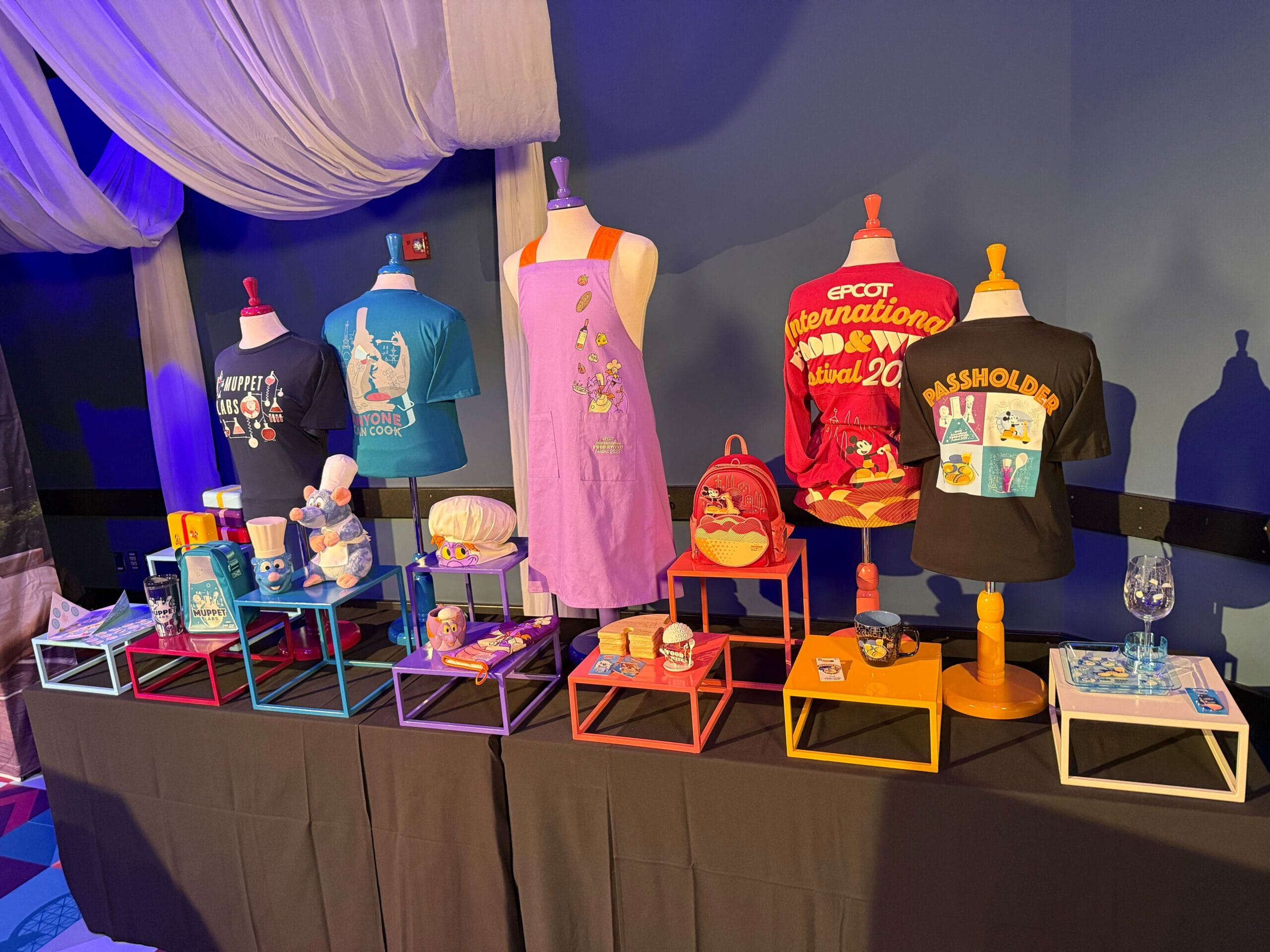 Colorful display of shirts, apron, mugs, and accessories on mannequins and tables at a merchandise booth.