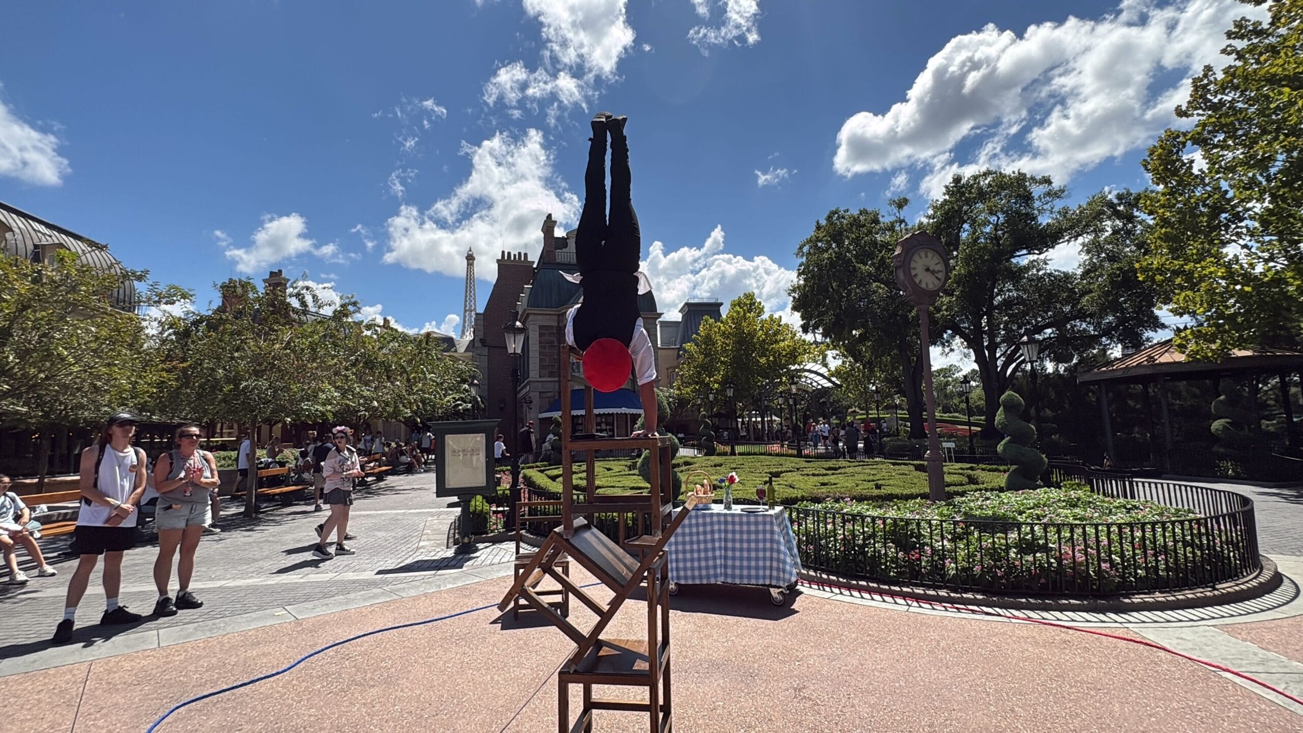 An acrobatic show at EPCOT features a street performer doing a handstand on stacked chairs as people watch in the park.