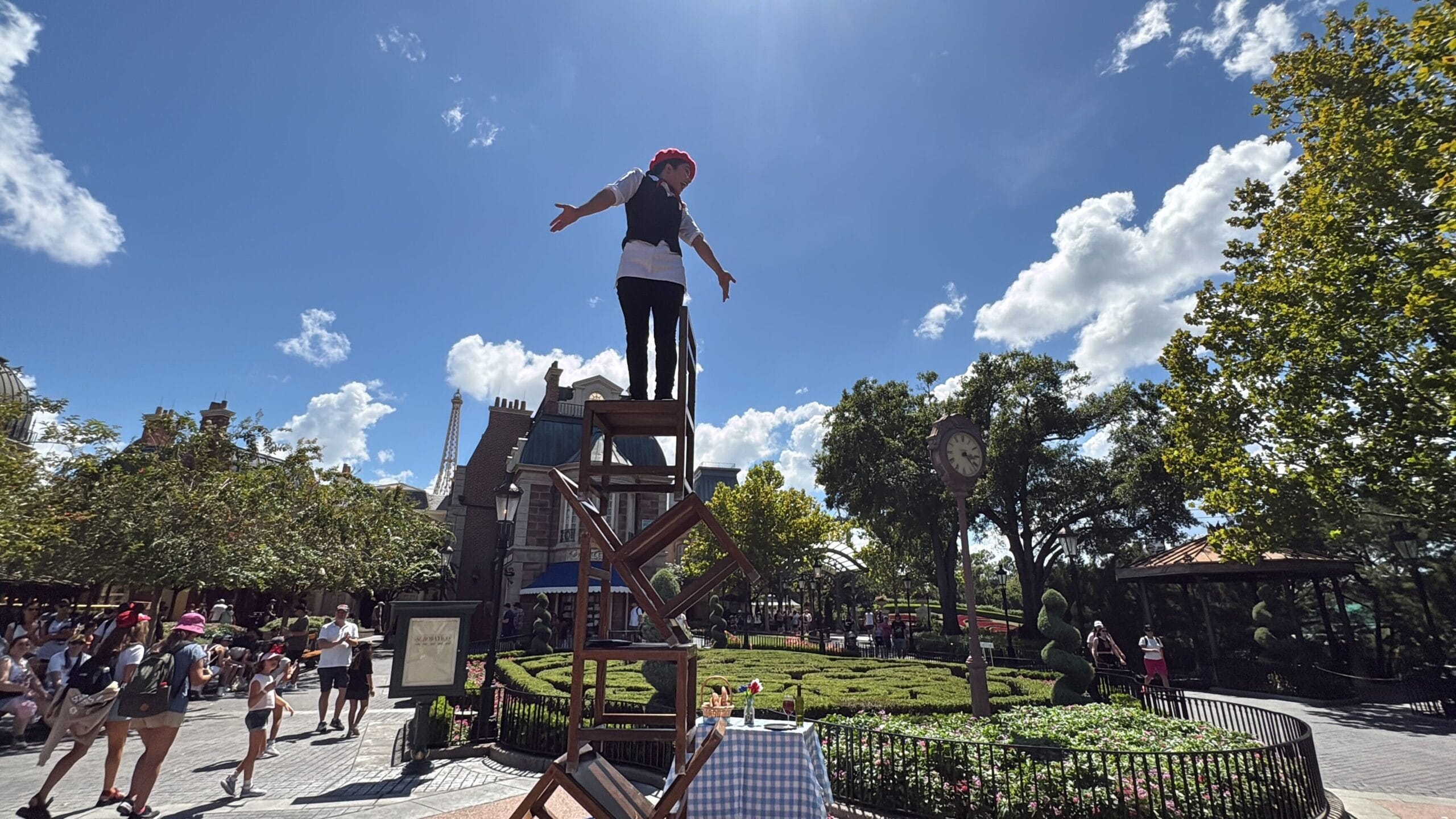 A performer stands with arms outstretched atop stacked chairs during an Acrobatico show in a sunny EPCOT park.