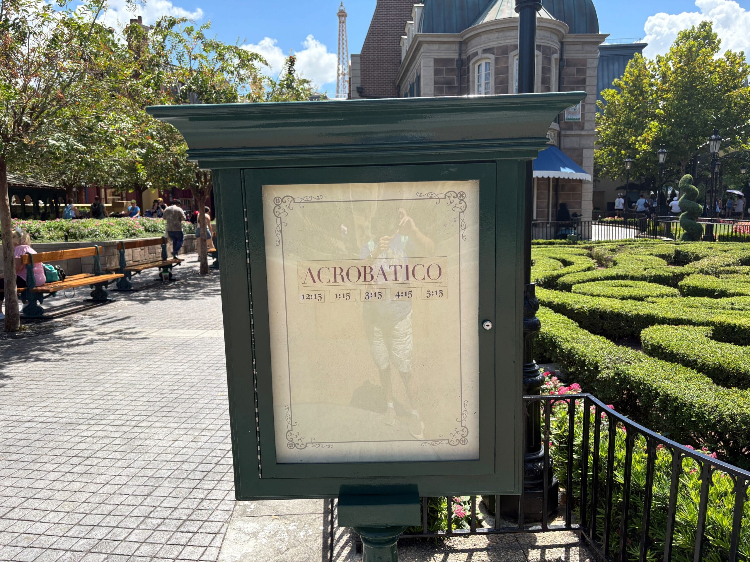 Sign reading "Acrobatico" with showtimes in a park at EPCOT, surrounded by greenery and people walking nearby.