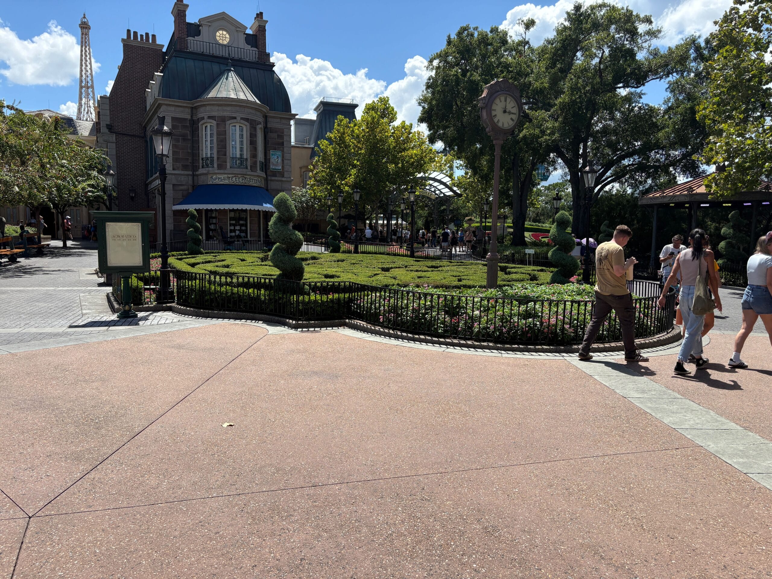 A landscaped garden with shaped topiary, a clock, and people walking near EPCOT’s European-style building under a blue sky.