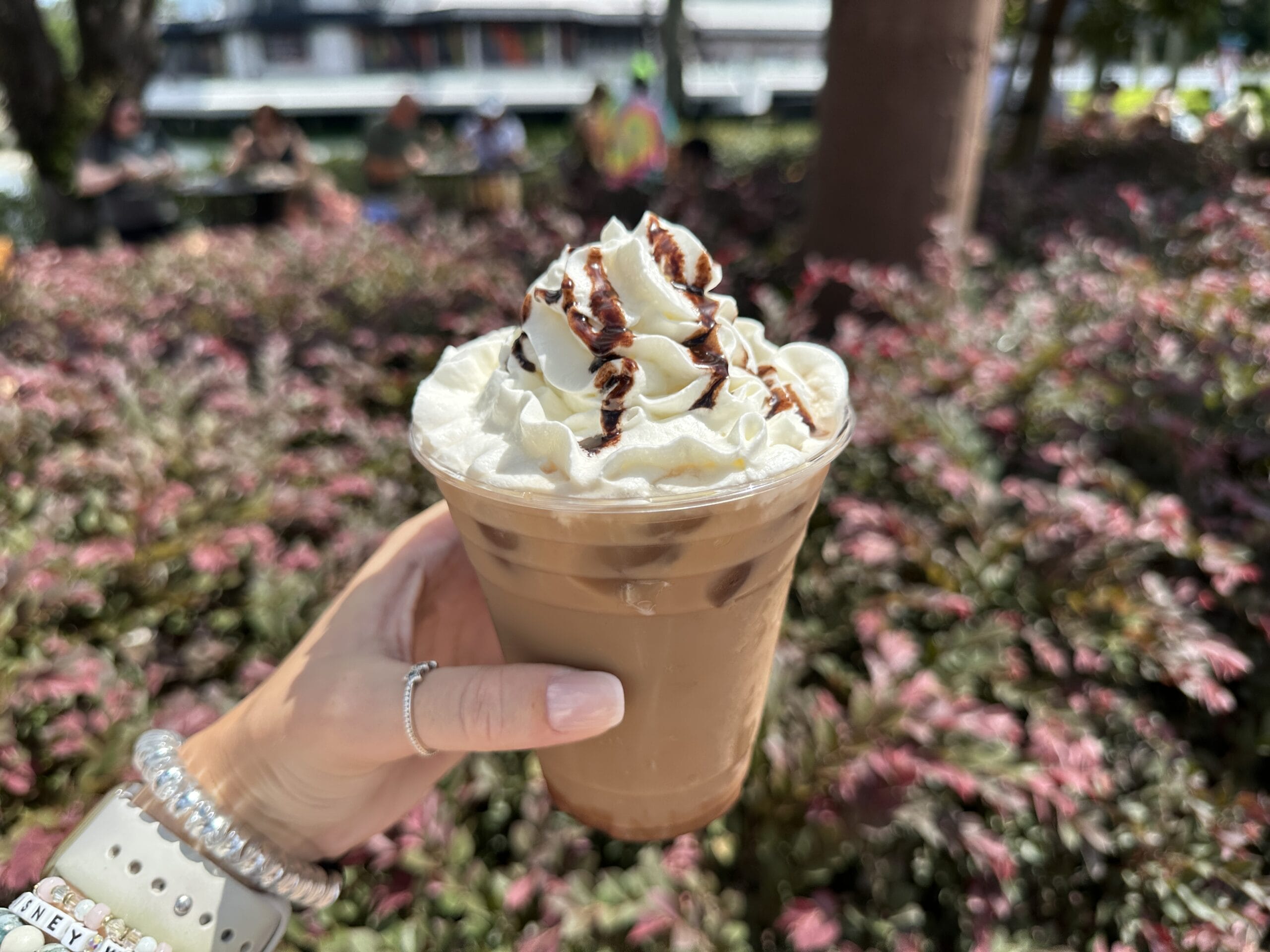 A hand holds an iced coffee topped with whipped cream and chocolate drizzle at the New Food & Wine event, near pink foliage.