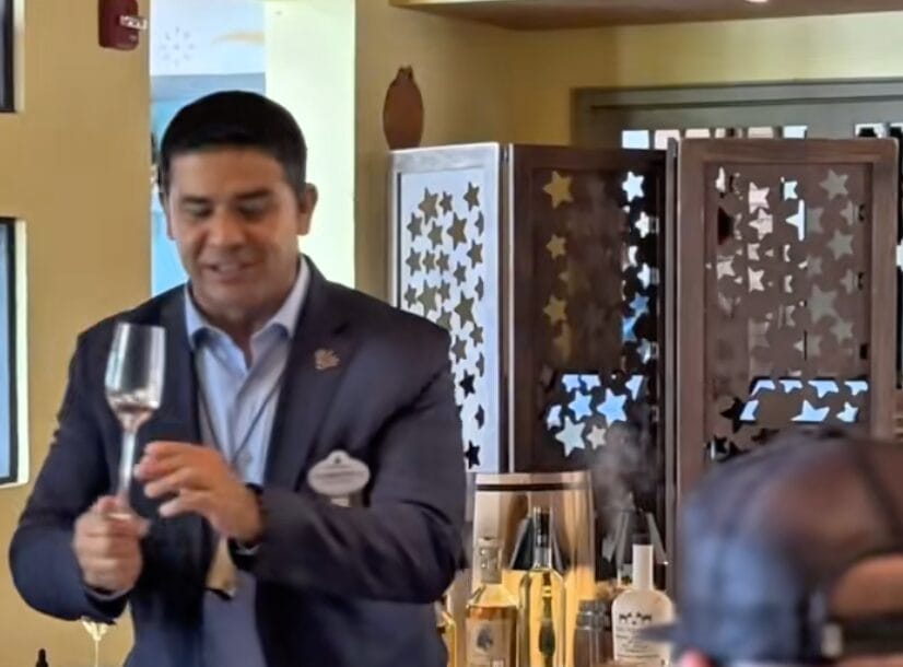 A man in a suit holds a wine glass in a bar area, with bottles and a decorative screen in the background.