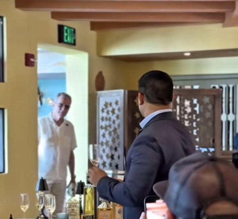 A man in white enters a room while another man in a suit holds a glass and looks toward him.