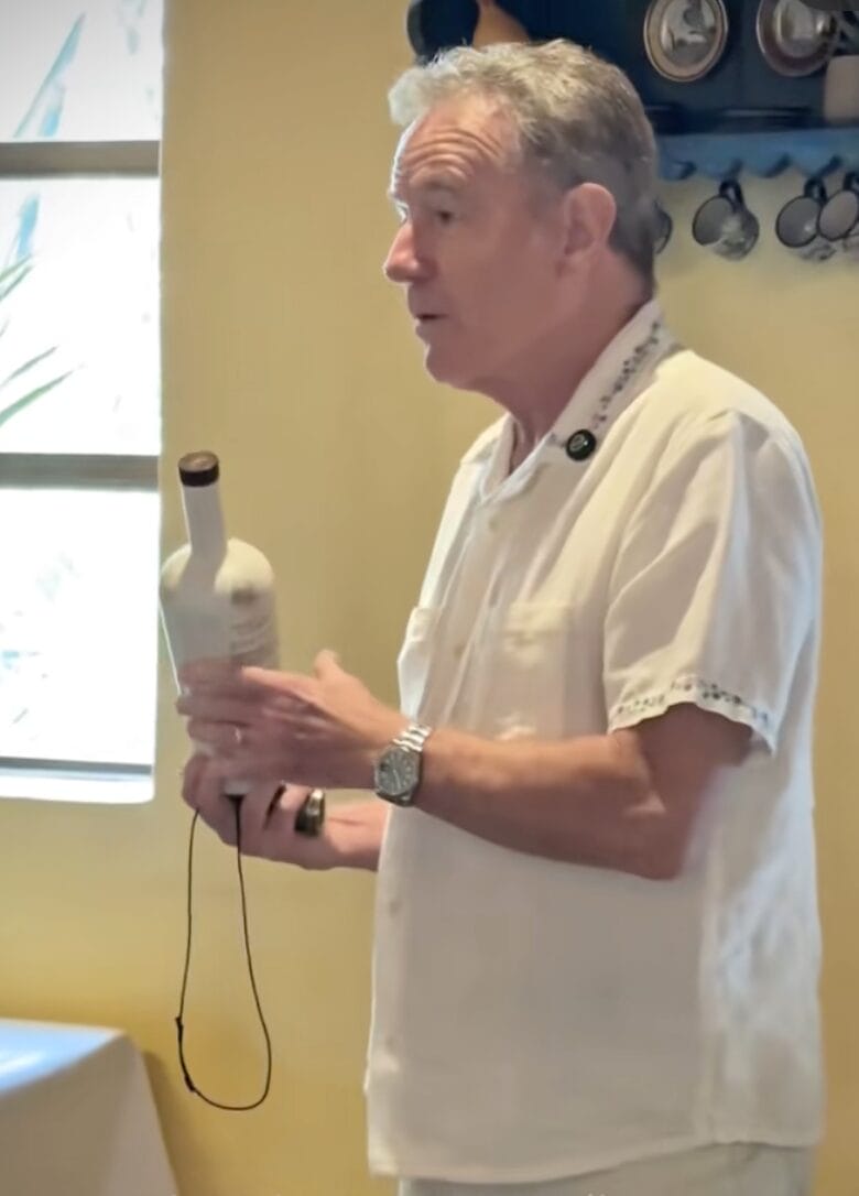 Bryan Cranston in a white shirt holds a bottle while speaking indoors near a window.