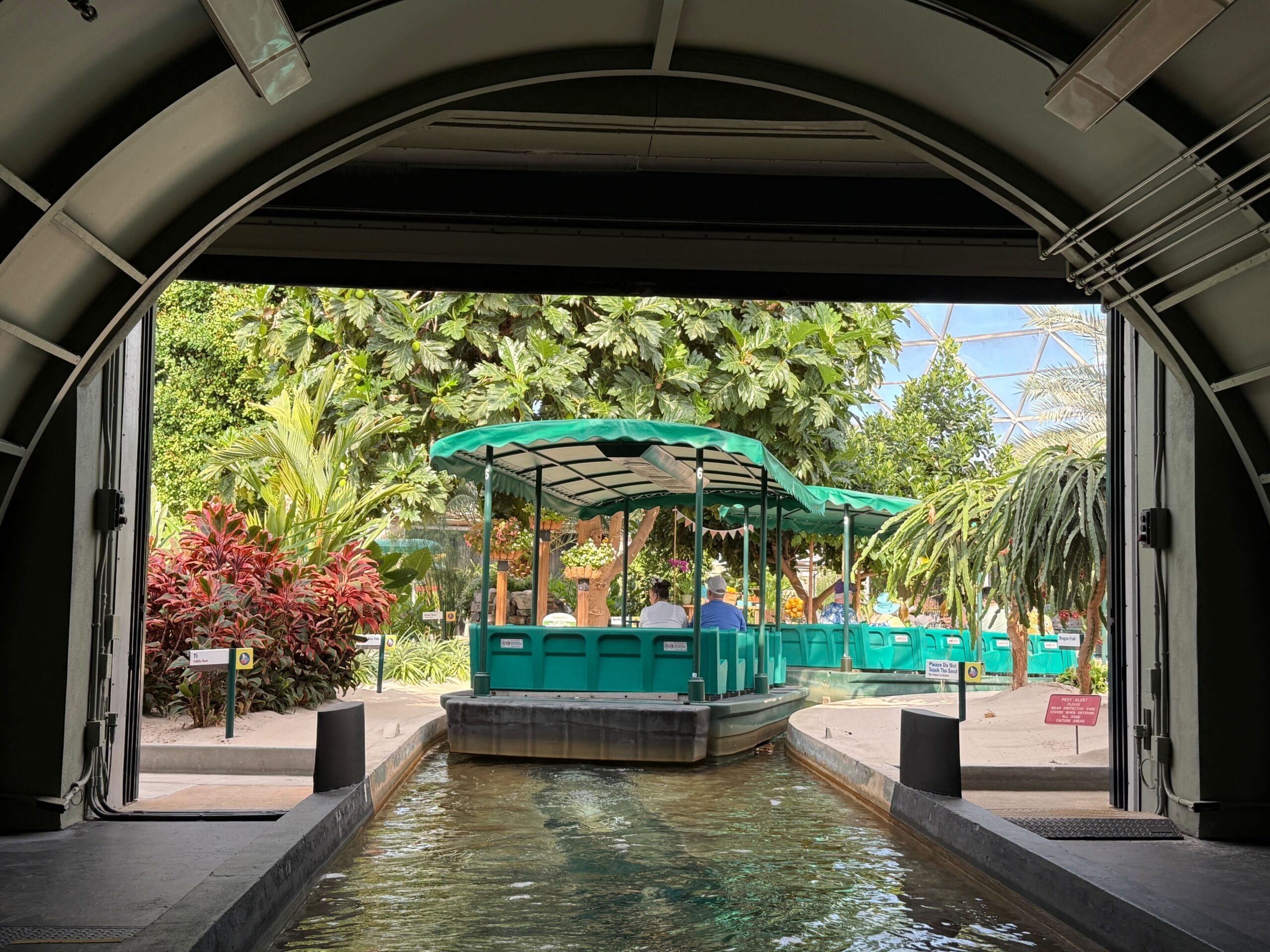A green boat ride on Living with the Land exits a tunnel into a lush, tropical garden with people under a canopy.