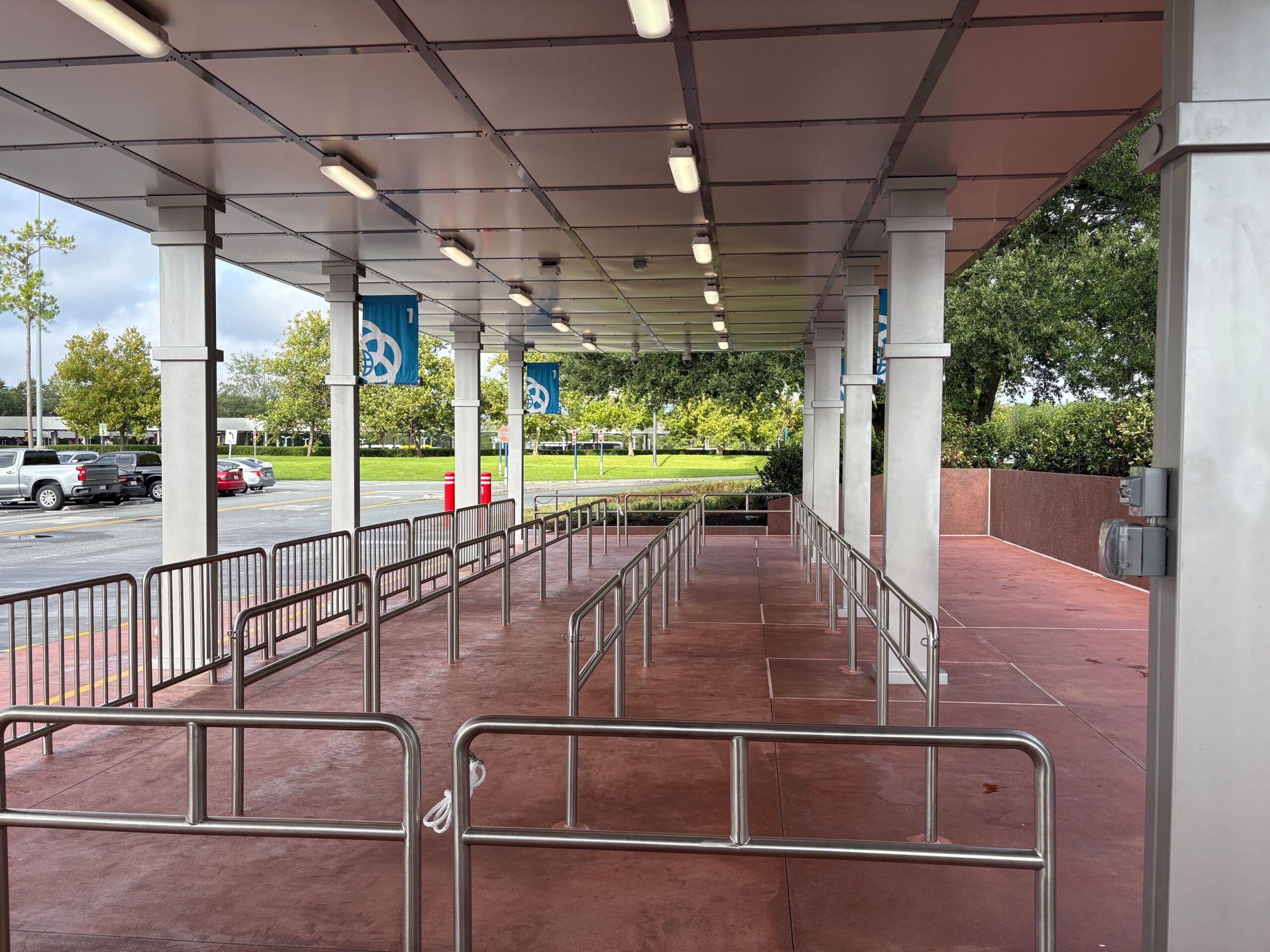 Empty queue area with metal railings under a covered walkway, next to a parking lot and green trees.