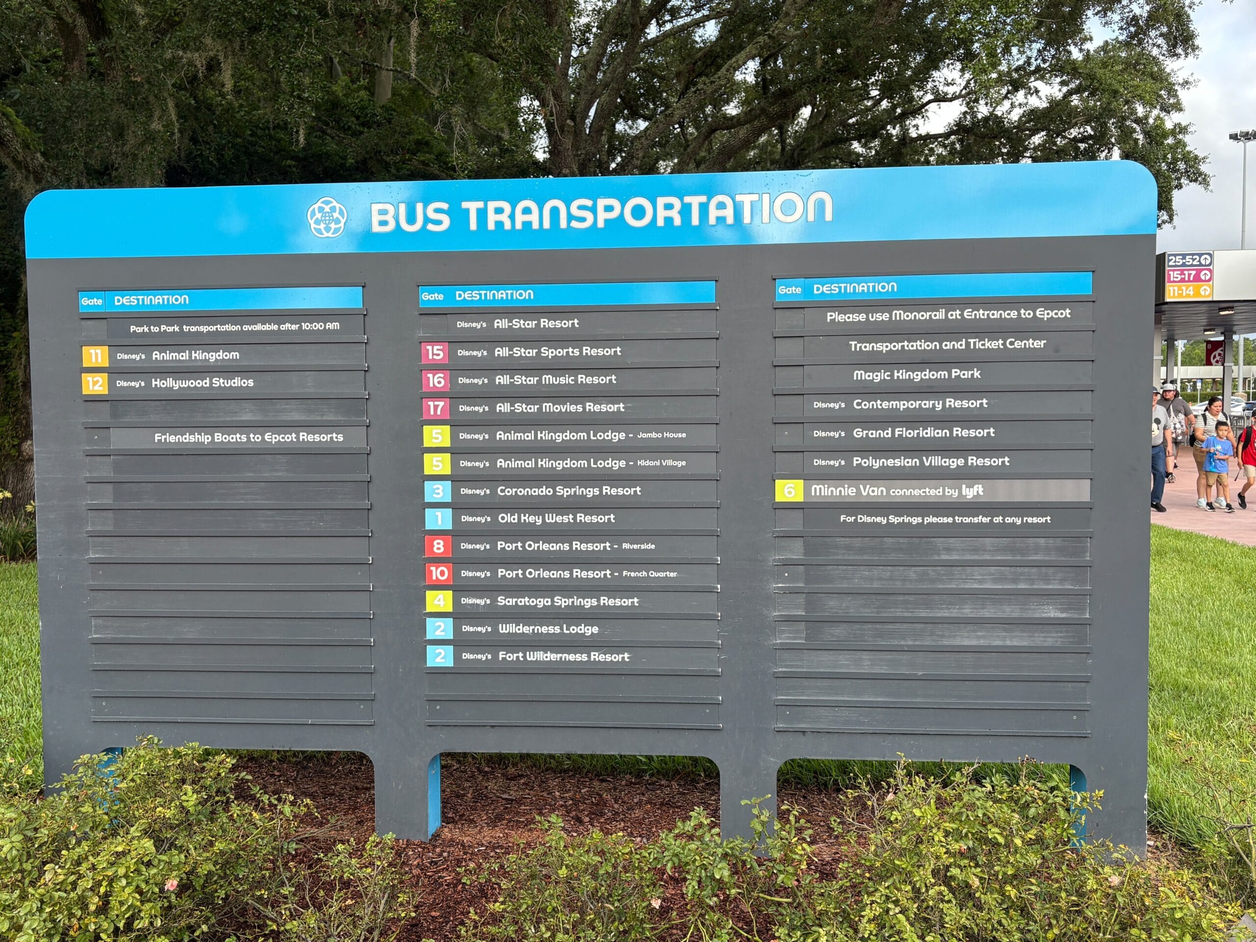 Disney World bus stop sign lists destinations like EPCOT, with trees and people visible in the background.