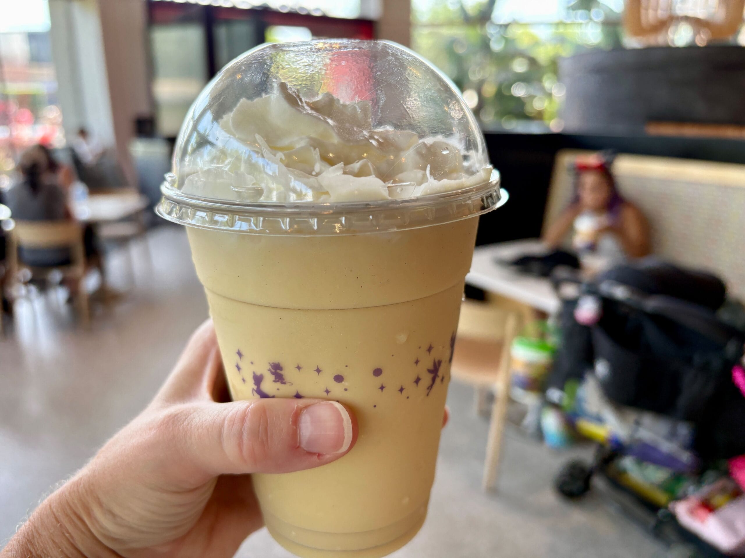 A hand holding a plastic cup of yellow milkshake topped with whipped cream, perfect for New Food & Wine lovers inside a cafe.