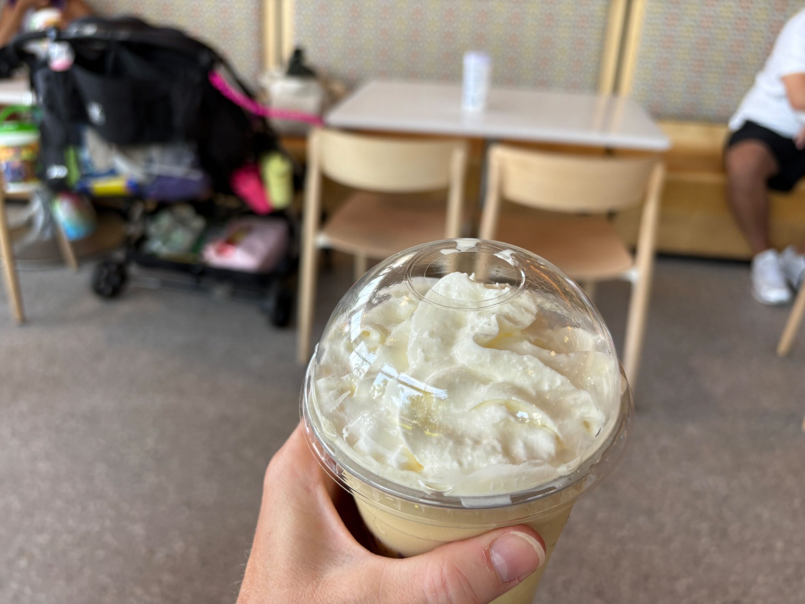 A hand holds a cold drink topped with whipped cream in a café, highlighting New Food & Wine trends.