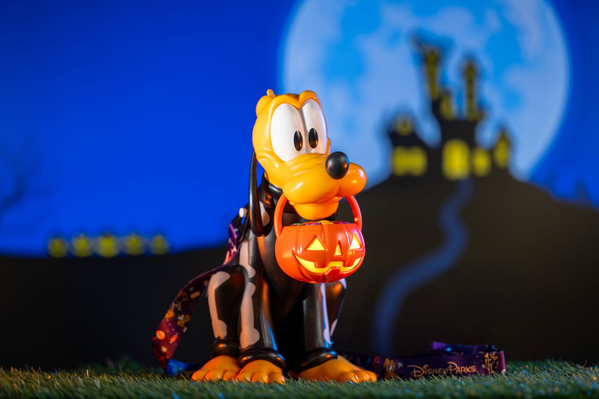 Pluto figurine wearing a cape holds a glowing jack-o’-lantern bucket, with a haunted house in the background.