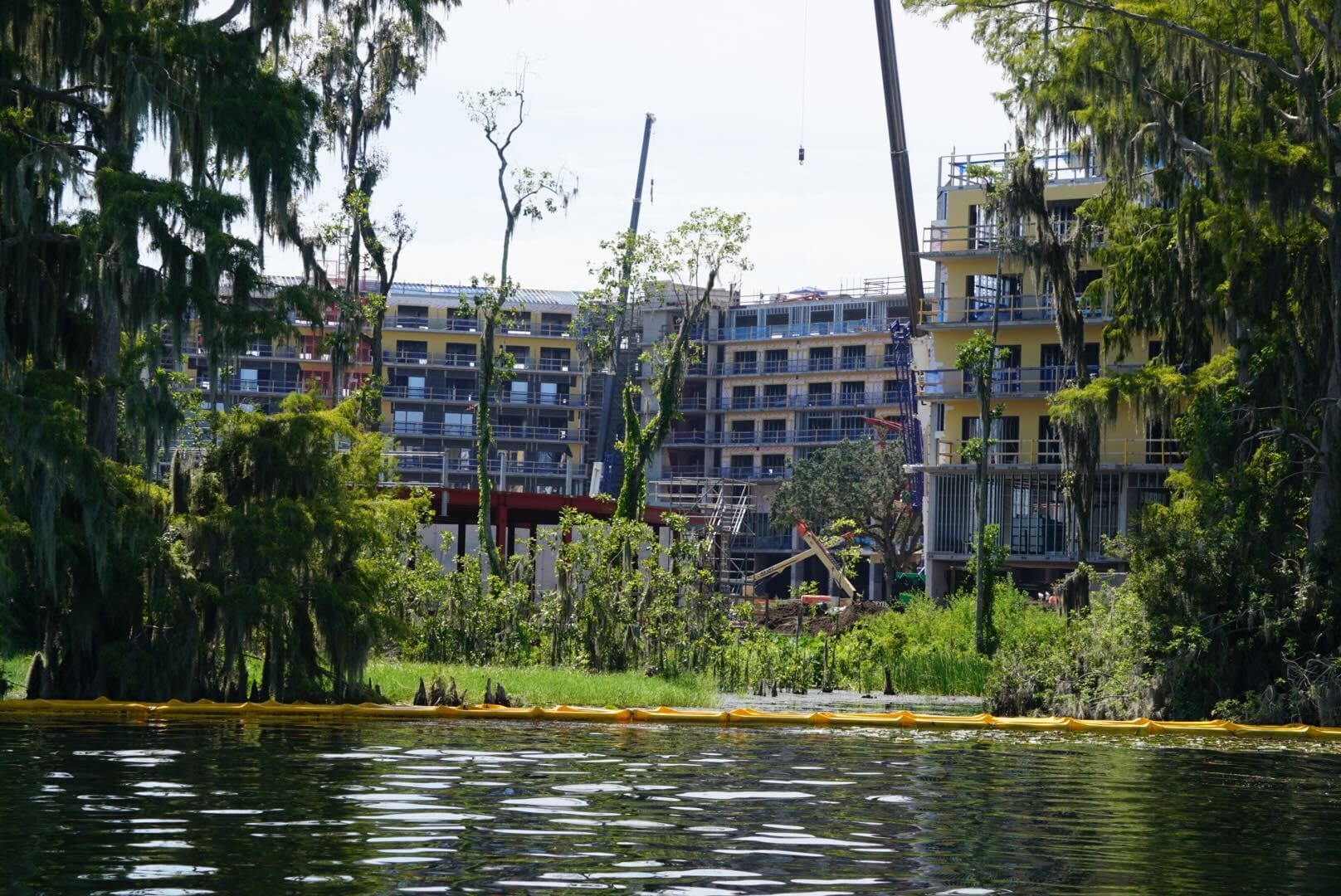 A large Lakeshore building under construction behind trees and a lake, with cranes and scaffolding visible.