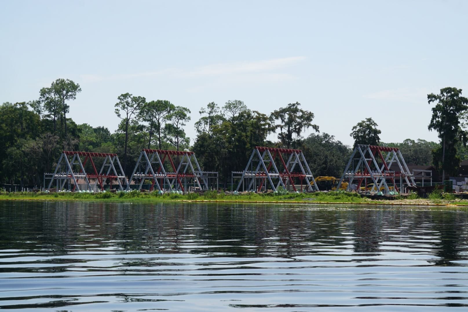 Four large metal A-frame structures rise along the lakeshore, reflected in calm water near a tree-lined shore.