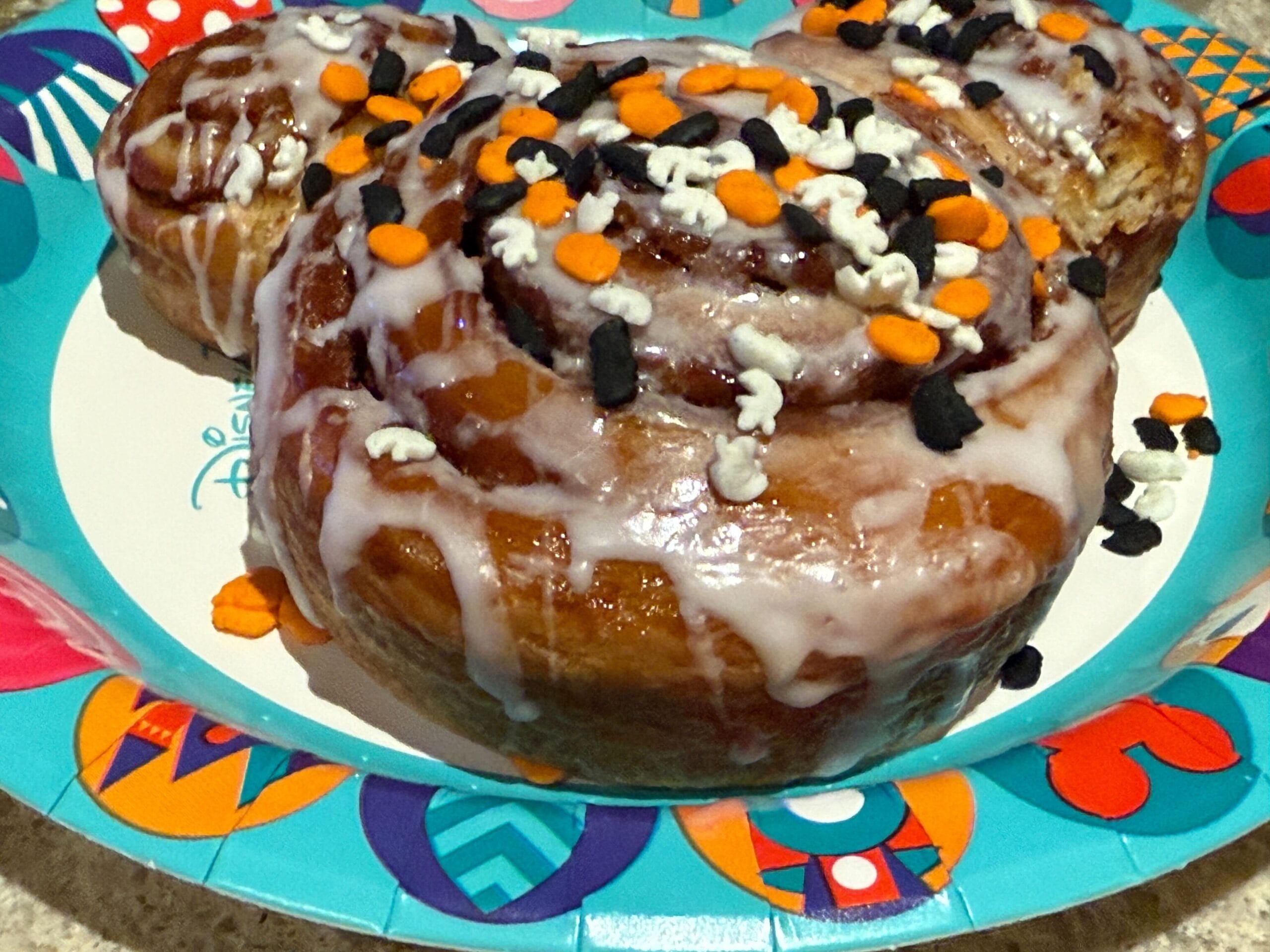 A Mickey Mouse-shaped cinnamon roll with icing and sprinkles from Mickey's Not So Scary Halloween Party on a colorful plate.