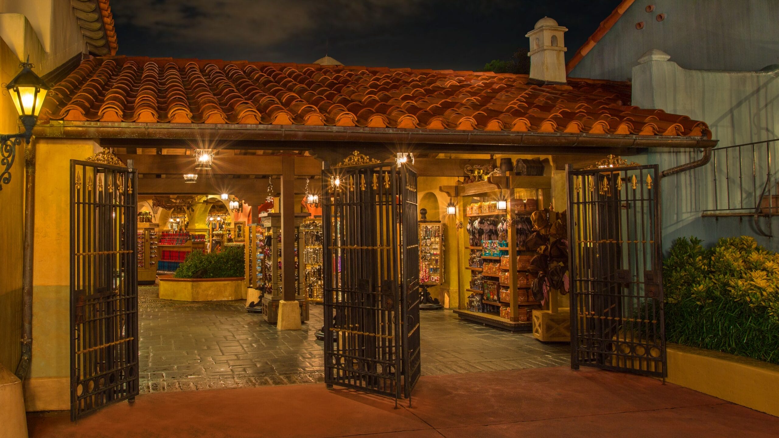 Open wrought-iron gates lead into Beak and Barrel Pirates Tavern, a warmly lit Disney World merchandise shop at night.