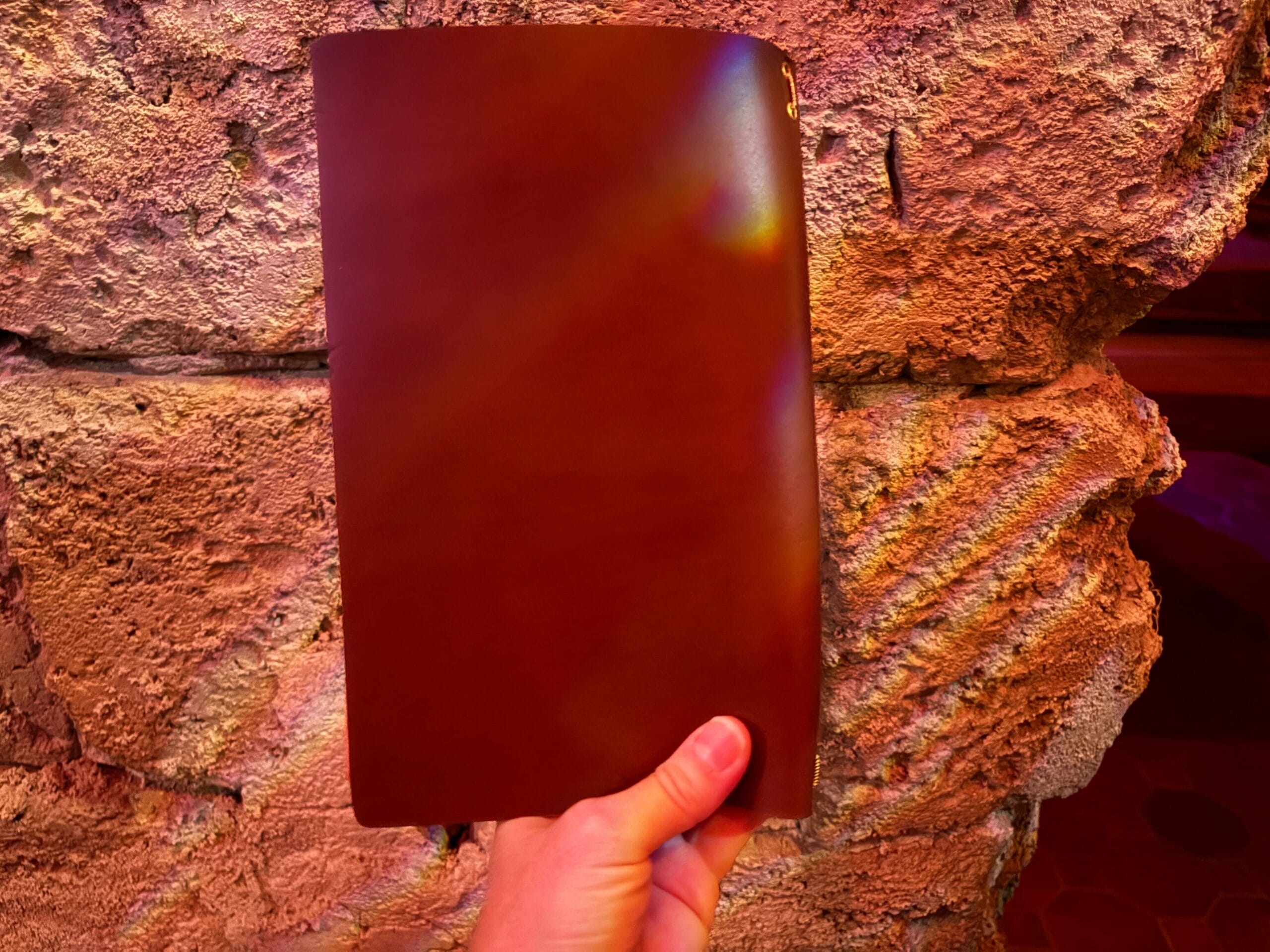 A hand holds a closed brown leather Beak and Barrel menu against a textured stone wall with warm lighting.