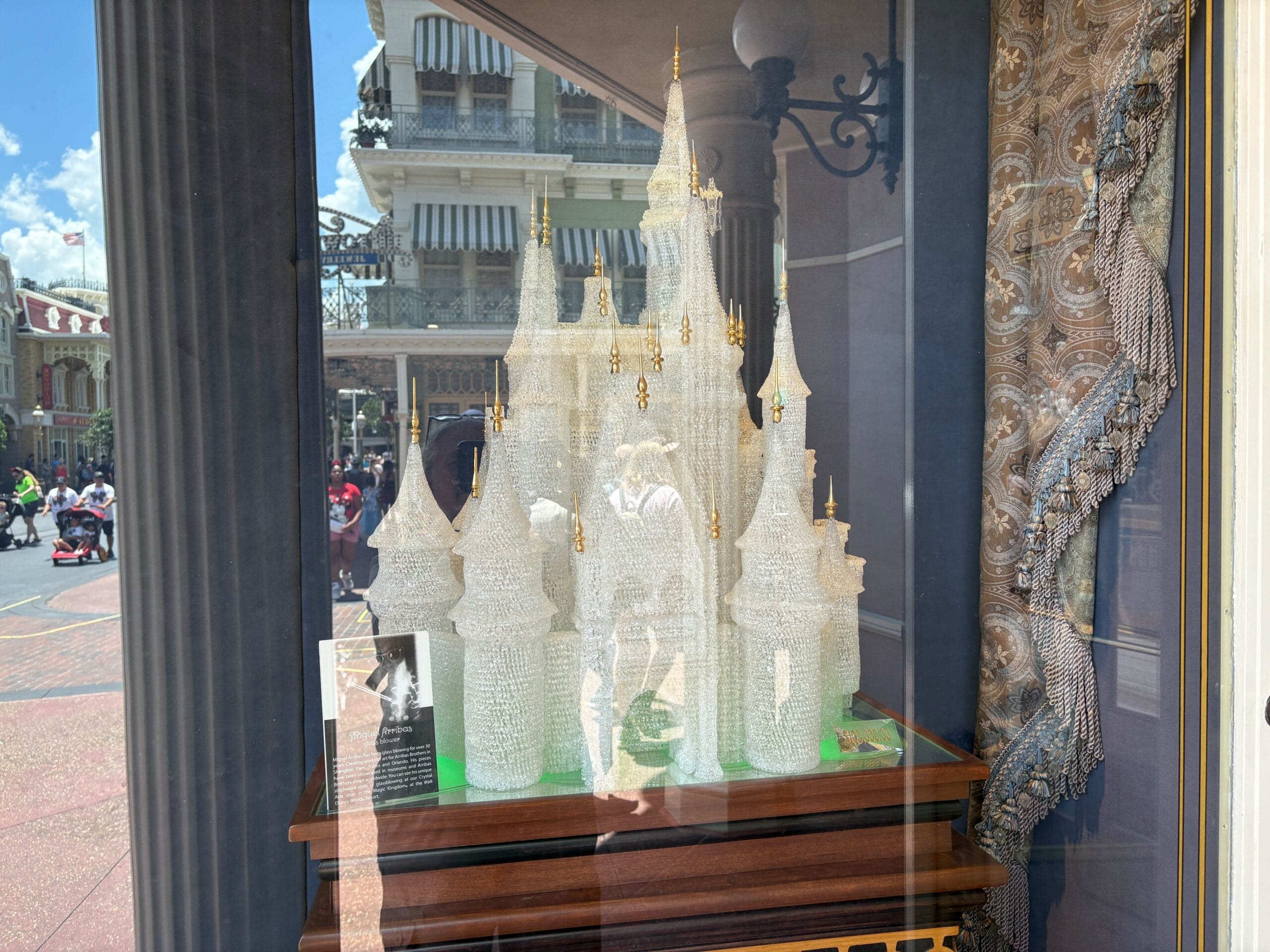 A crystal castle sculpture is displayed in a window with reflections of people and buildings outside.