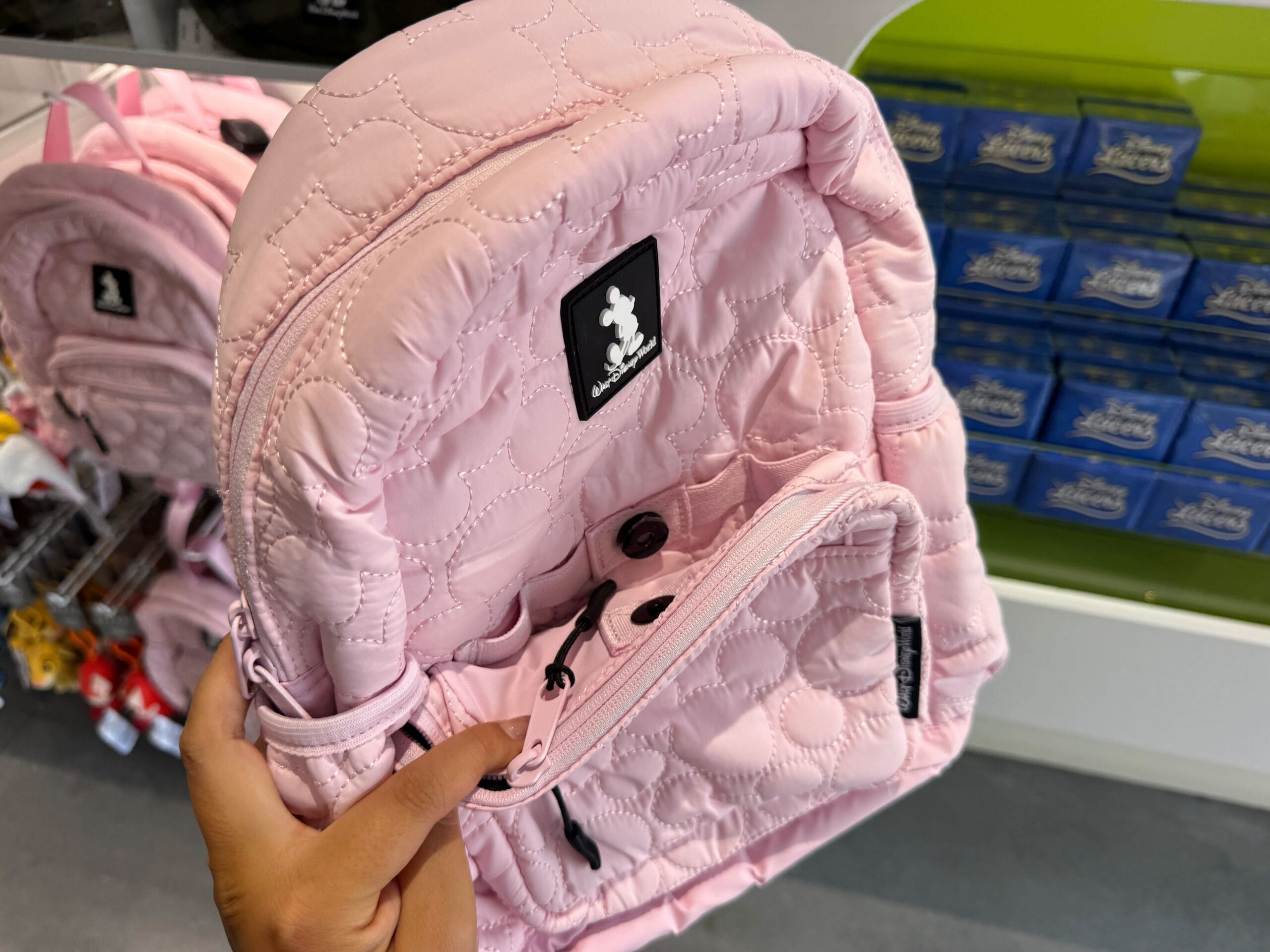 A hand holds a light pink Quilted Mickey Mouse Backpack with a black logo patch in a retail store.